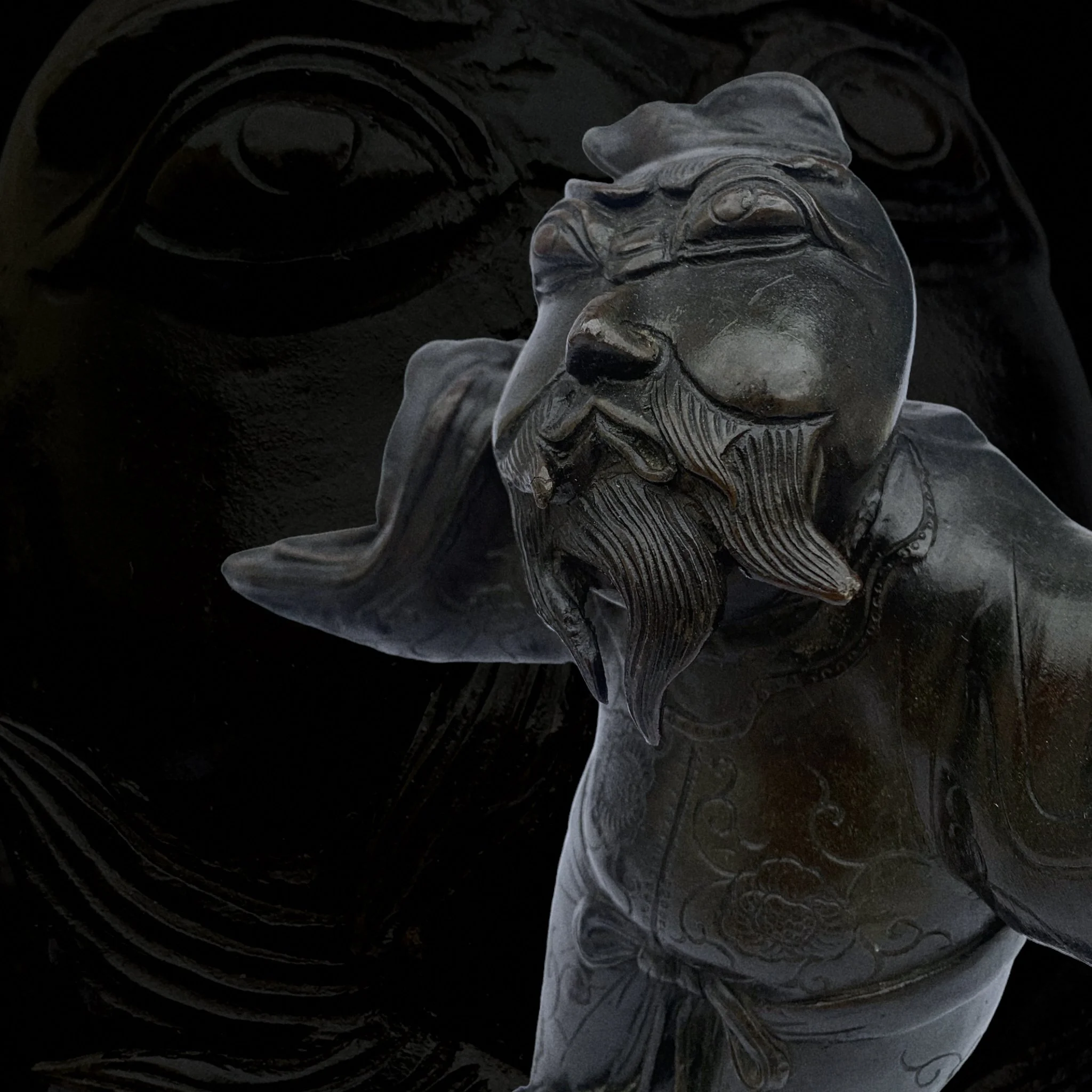 Dark-colored sculpture of a bearded man with a serious expression, detailed carvings on his clothing, and an elaborate style, set against a similarly dark background.