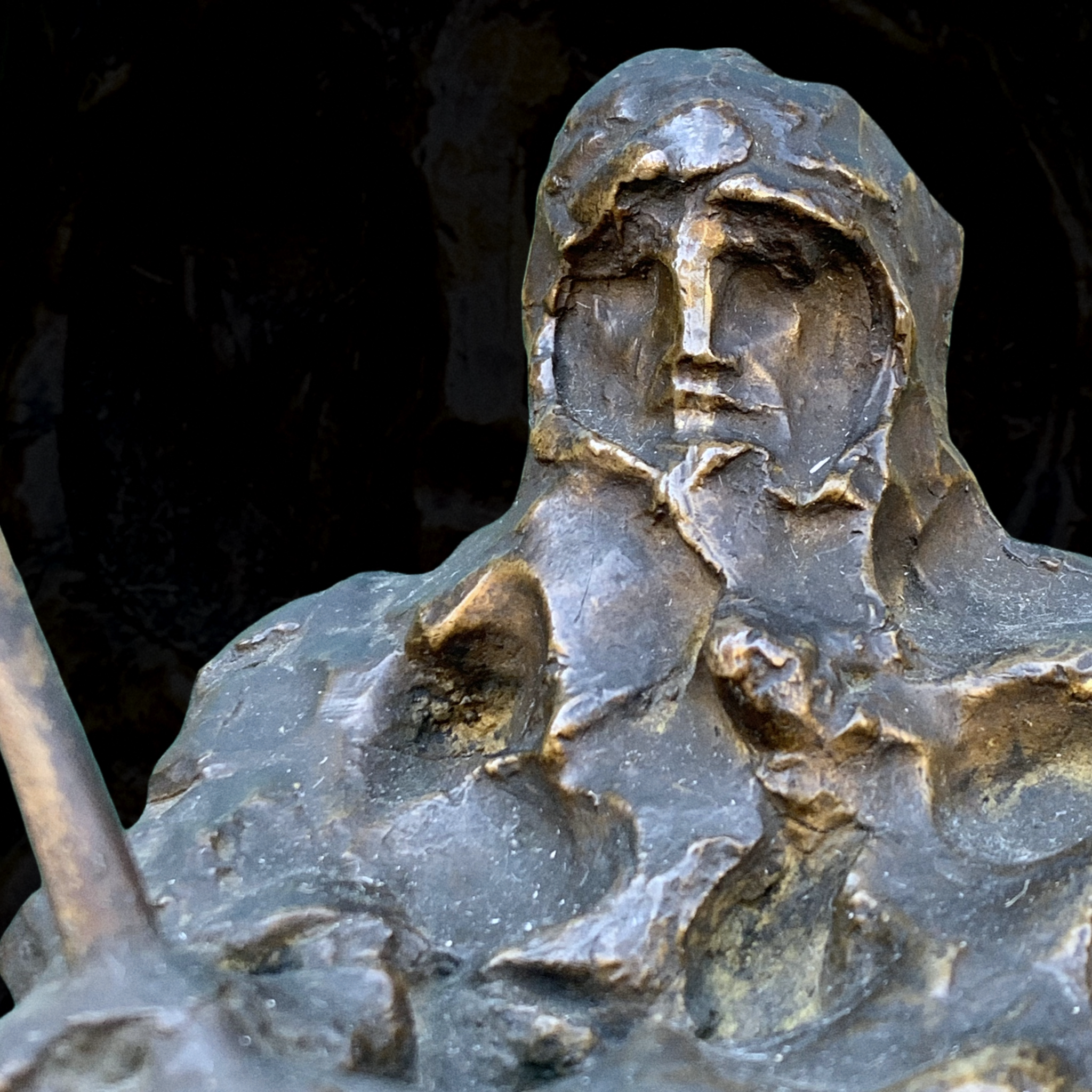 Close-up of a bronze sculpture of a woman with a serious expression, portrayed with flowing hair and a cloak, set against a dark background.