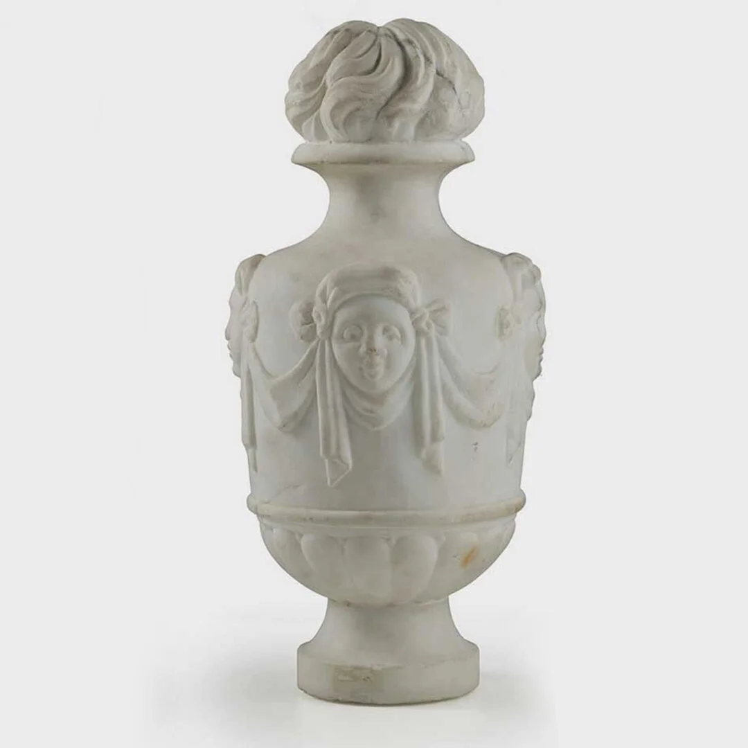 A white marble decorative vase with carved faces and detailed hair and clothing accents.
