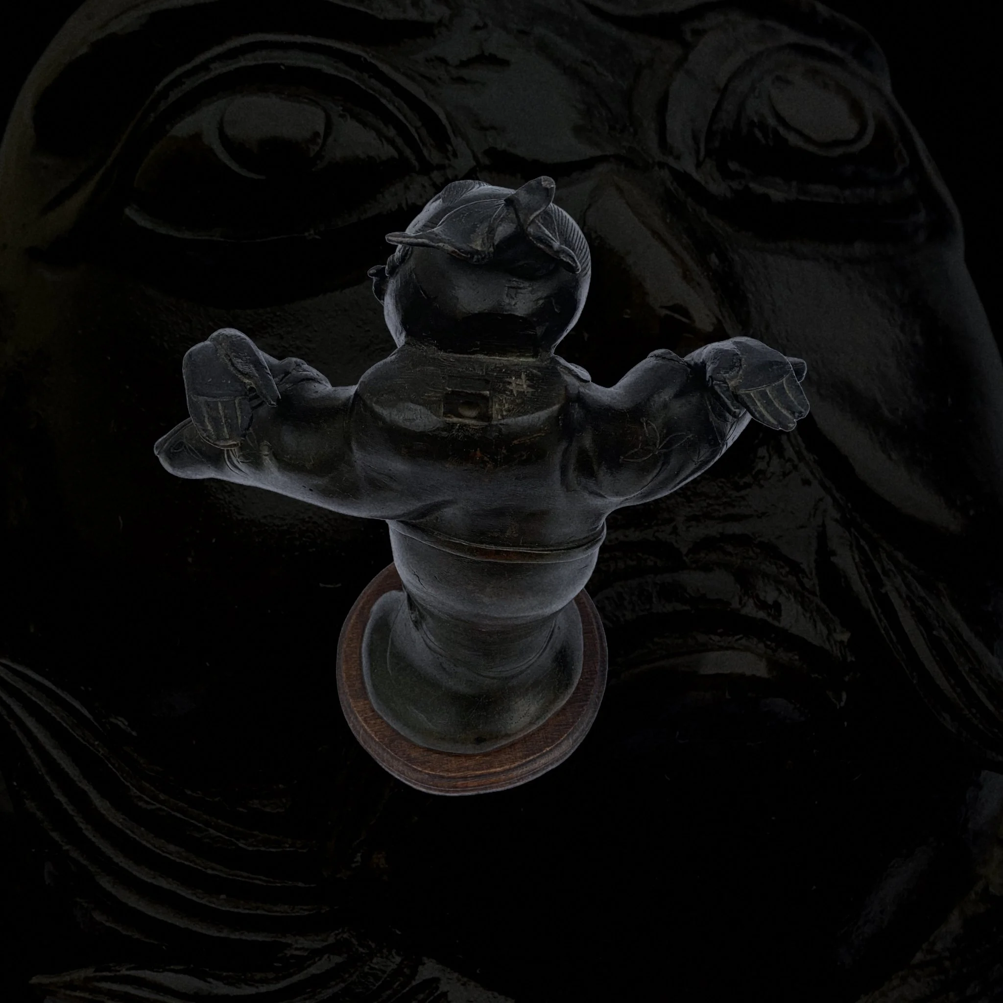 A sculpture of a cartoonish character with a round head, big eyes, and a wide smile, standing with arms open. The sculpture appears to be made of dark material and is displayed on a round base.