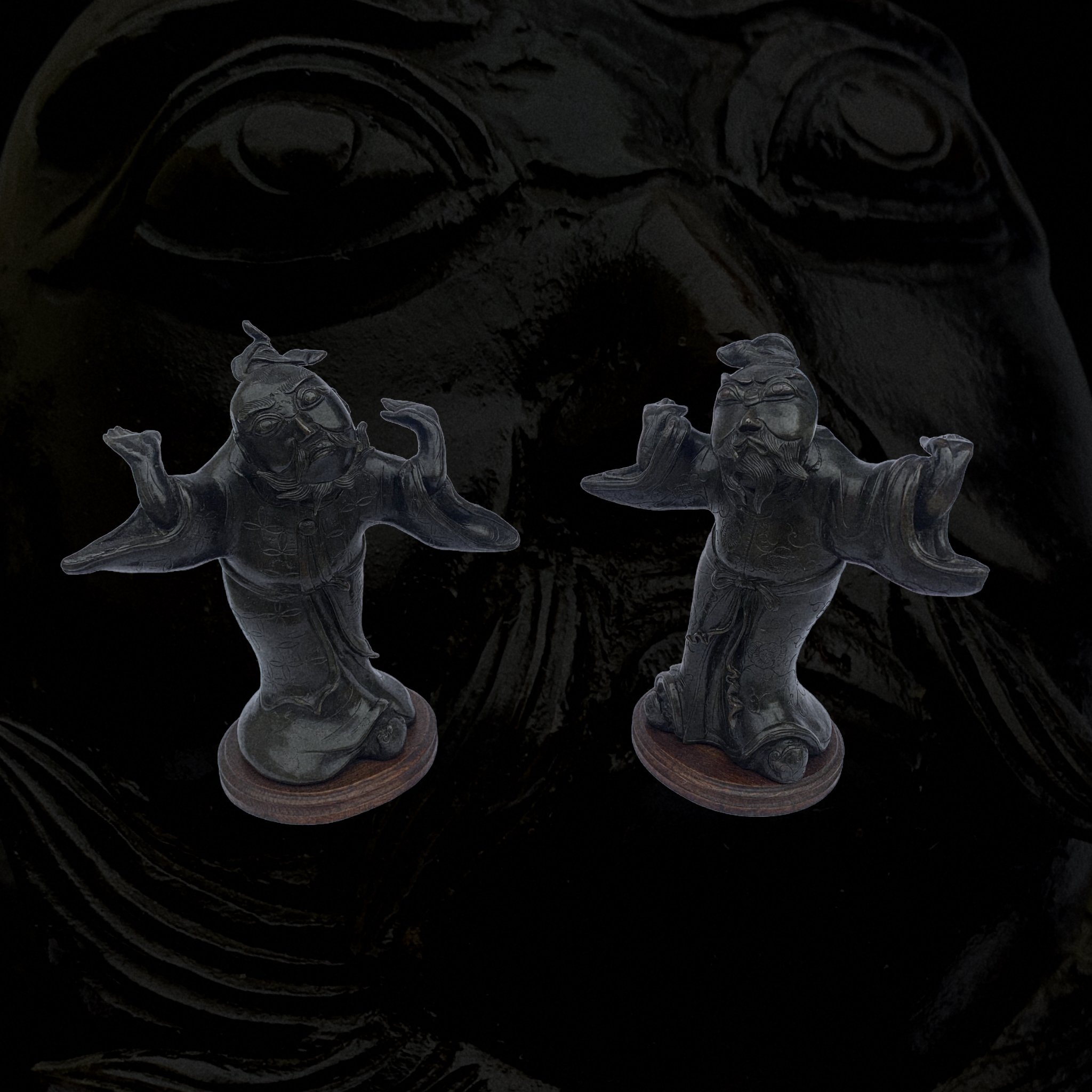 Two small, dark figurines of laughing, dressed figures with facial hair, on round bases, placed against a dark background.