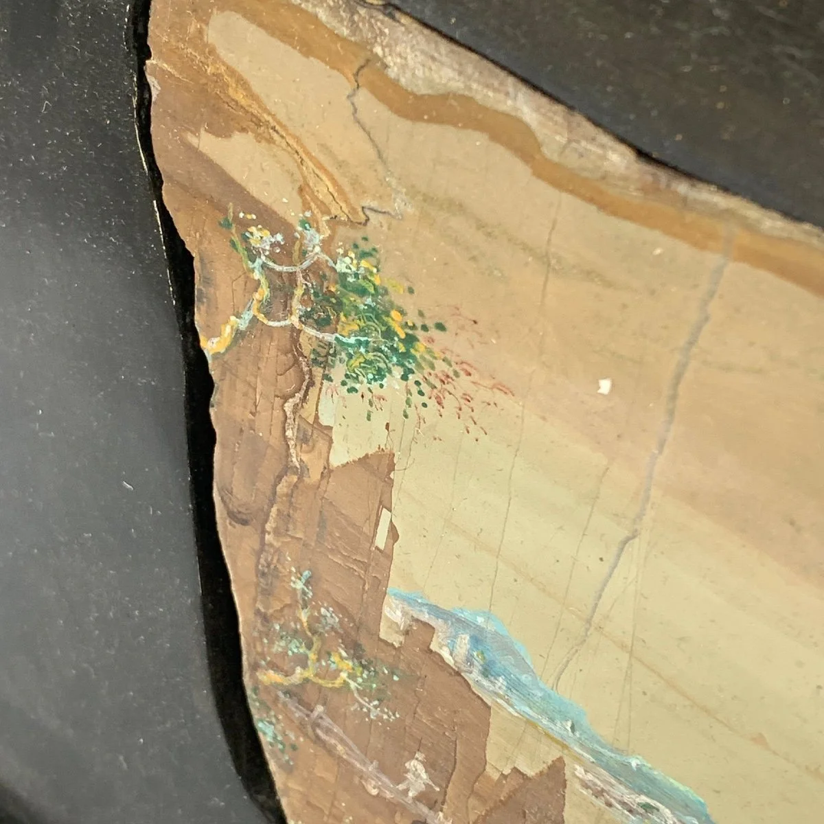 Close-up of a cracked and peeling painted surface with a small painted tree and landscape scene, partially obscured by black framing on the left side.