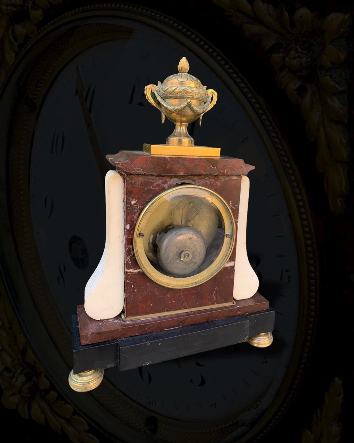 An antique clock with a red marble and white ceramic body, featuring a brass circular face and a decorative urn on top, displayed in front of a dark, ornate background.