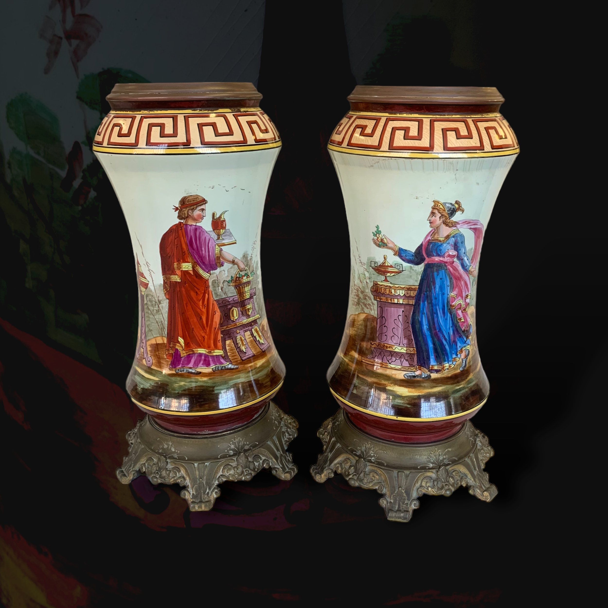 Two ornate vases with hand-painted scenes of classical women in colorful garments, each standing on decorative bases, set against a dark background.