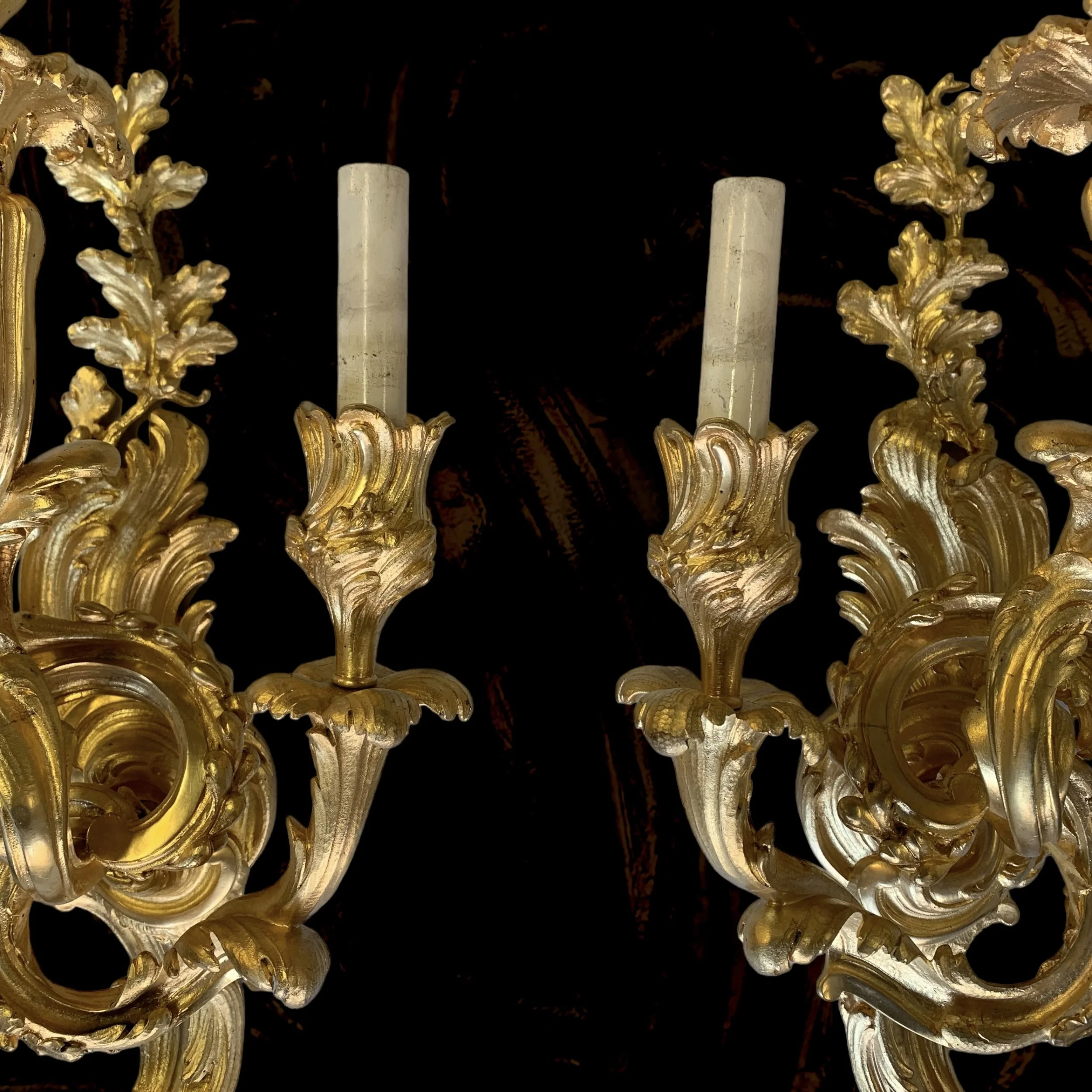 Close-up of a gold ornate chandelier with two white candle-like light fixtures against a dark background.