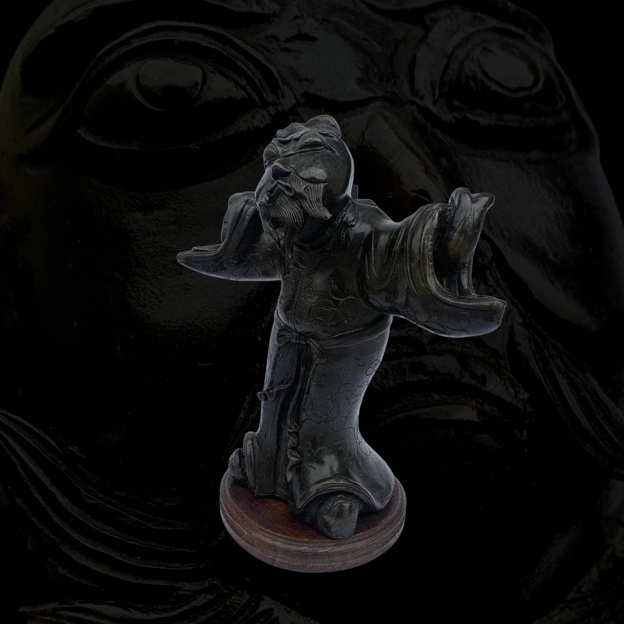 A carved Asian warrior figure with a fierce expression, standing on a round wooden base, set against a dark background.