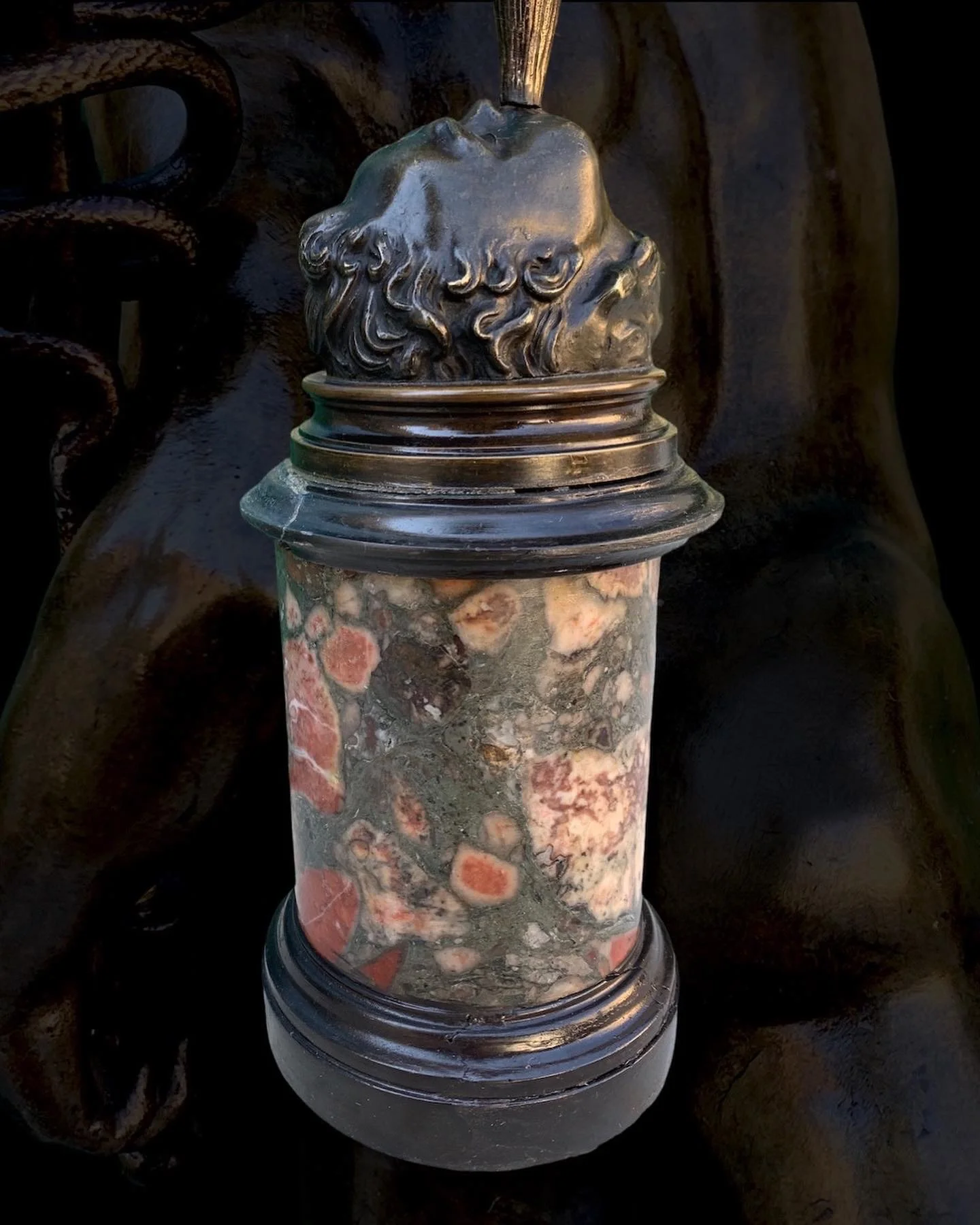 A decorative glass or stone cylindrical object with a bronze-colored, ornate lion's head top, set against a dark background.