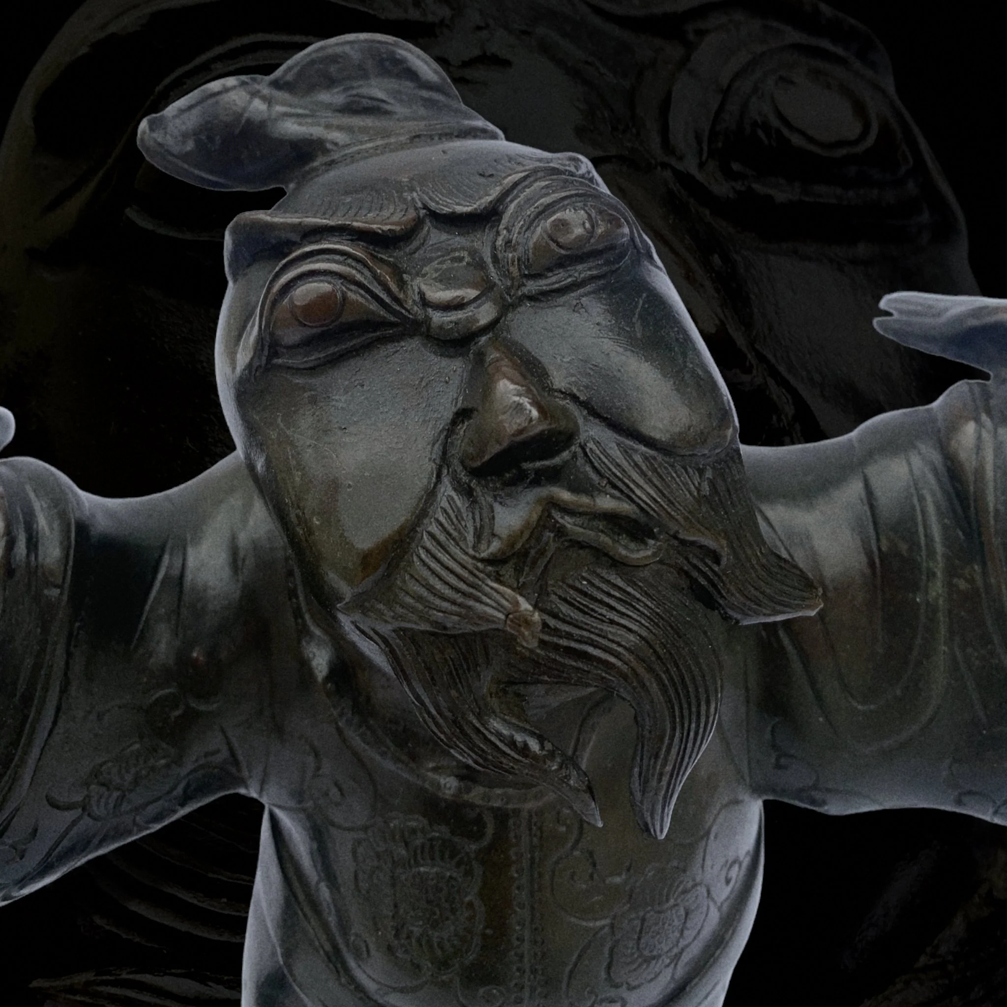 Close-up of a dark-colored, intricately carved sculpture of a figure with an expressive face, beard, and headpiece, set against a black background.