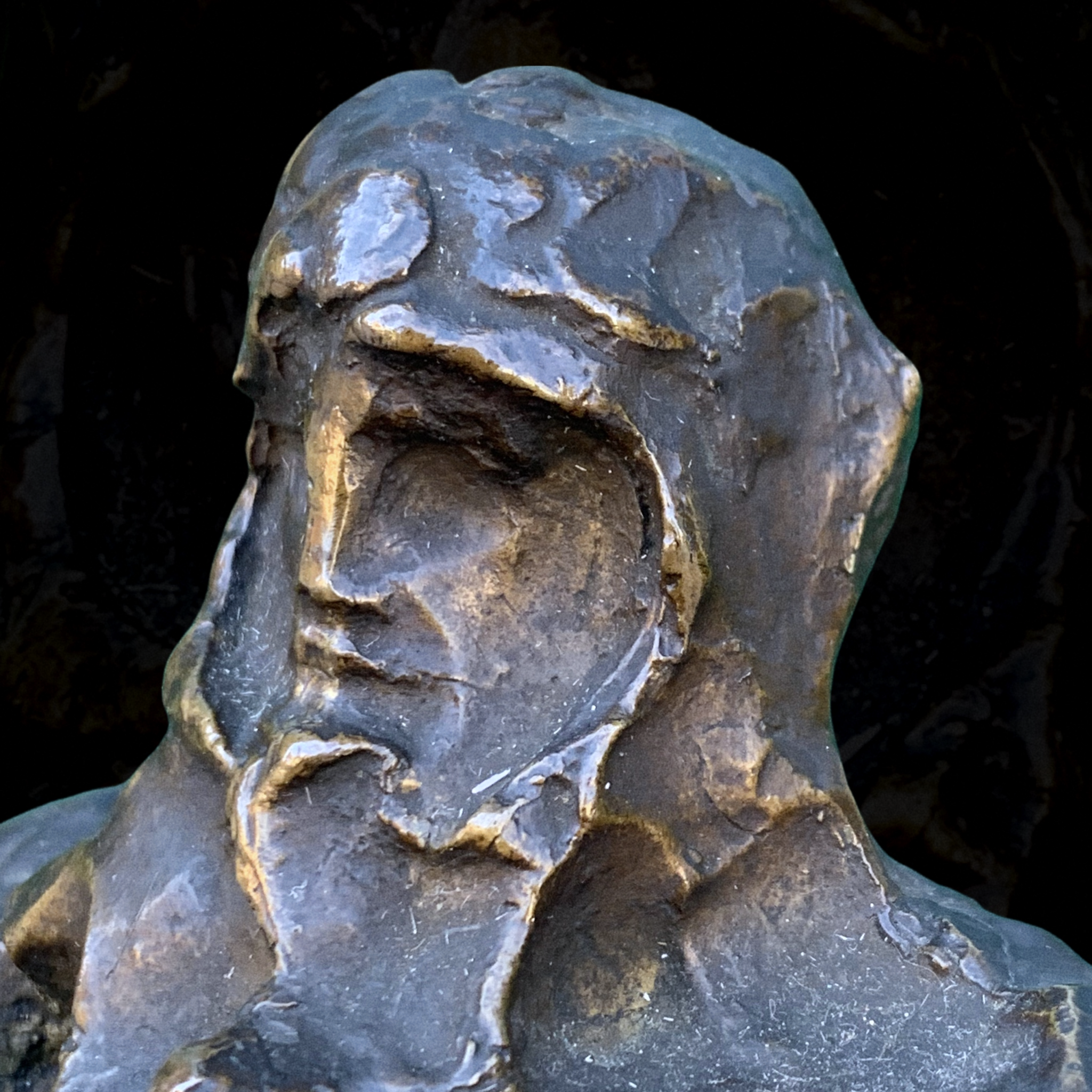 Close-up of a weathered bronze sculpture of a person with a beard and mustache, looking to the left, with a dark background.
