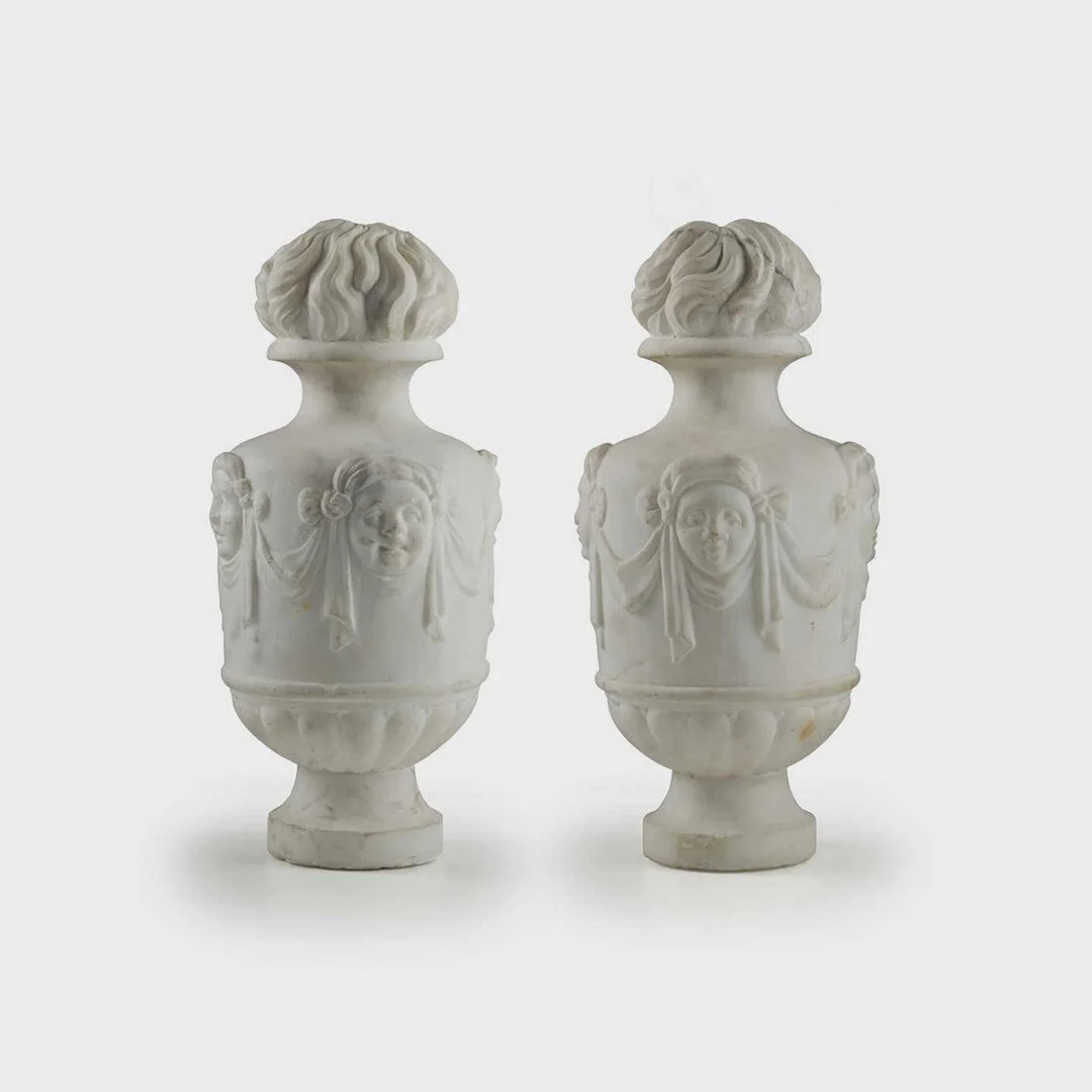 Two white marble decorative vases with carved faces and draped figure details, set against a plain background.
