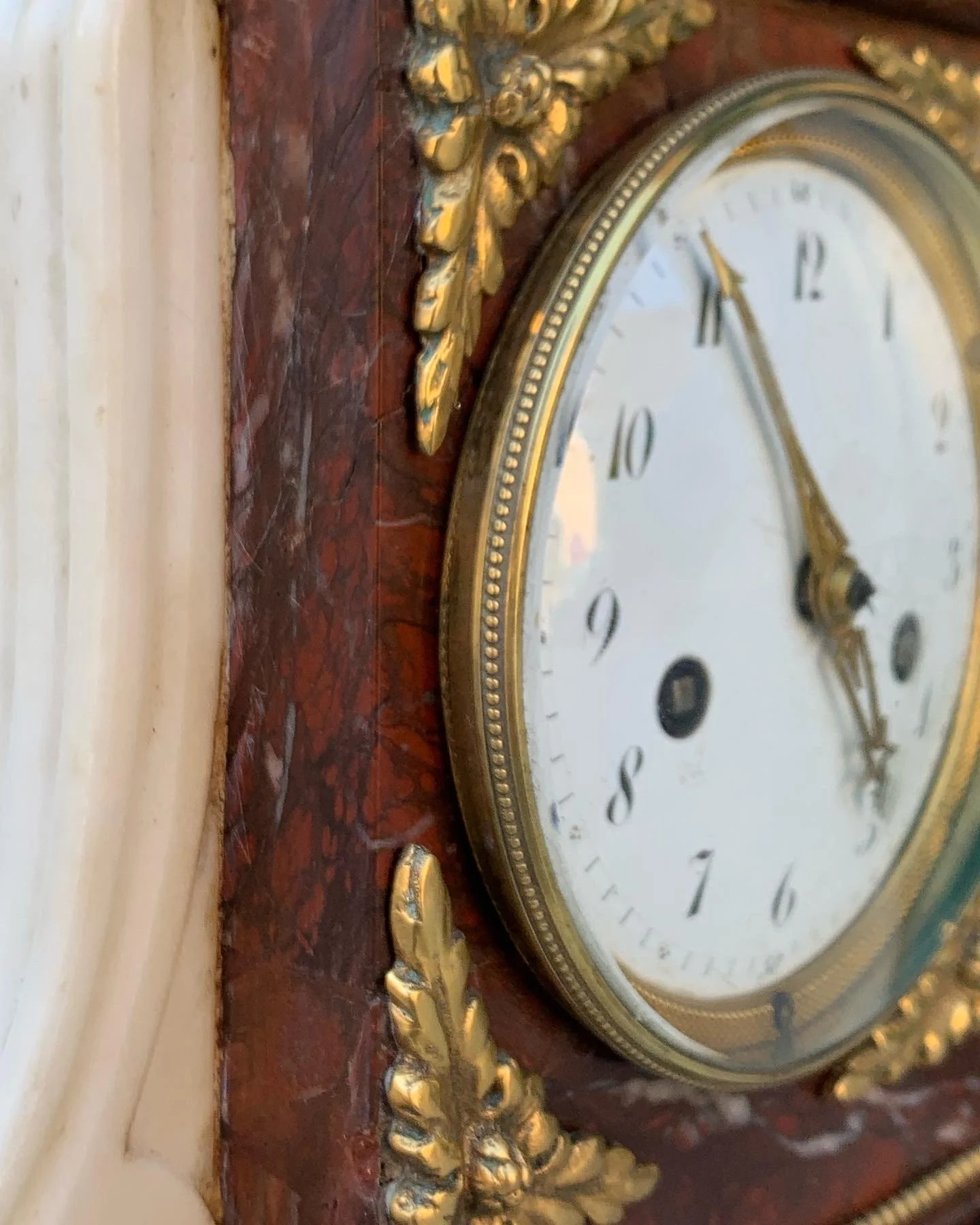 An antique clock with a white face and black numbers, encased in a gold and glass frame, mounted on a red marble wall with ornate gold decoration.