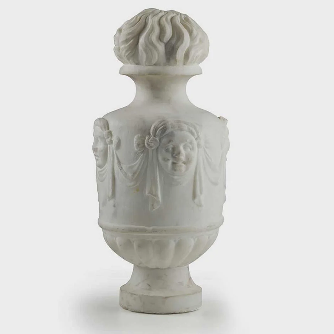 White marble vase with sculpted faces and draped fabric reliefs, topped with a decorative lid resembling a flower or fruit.