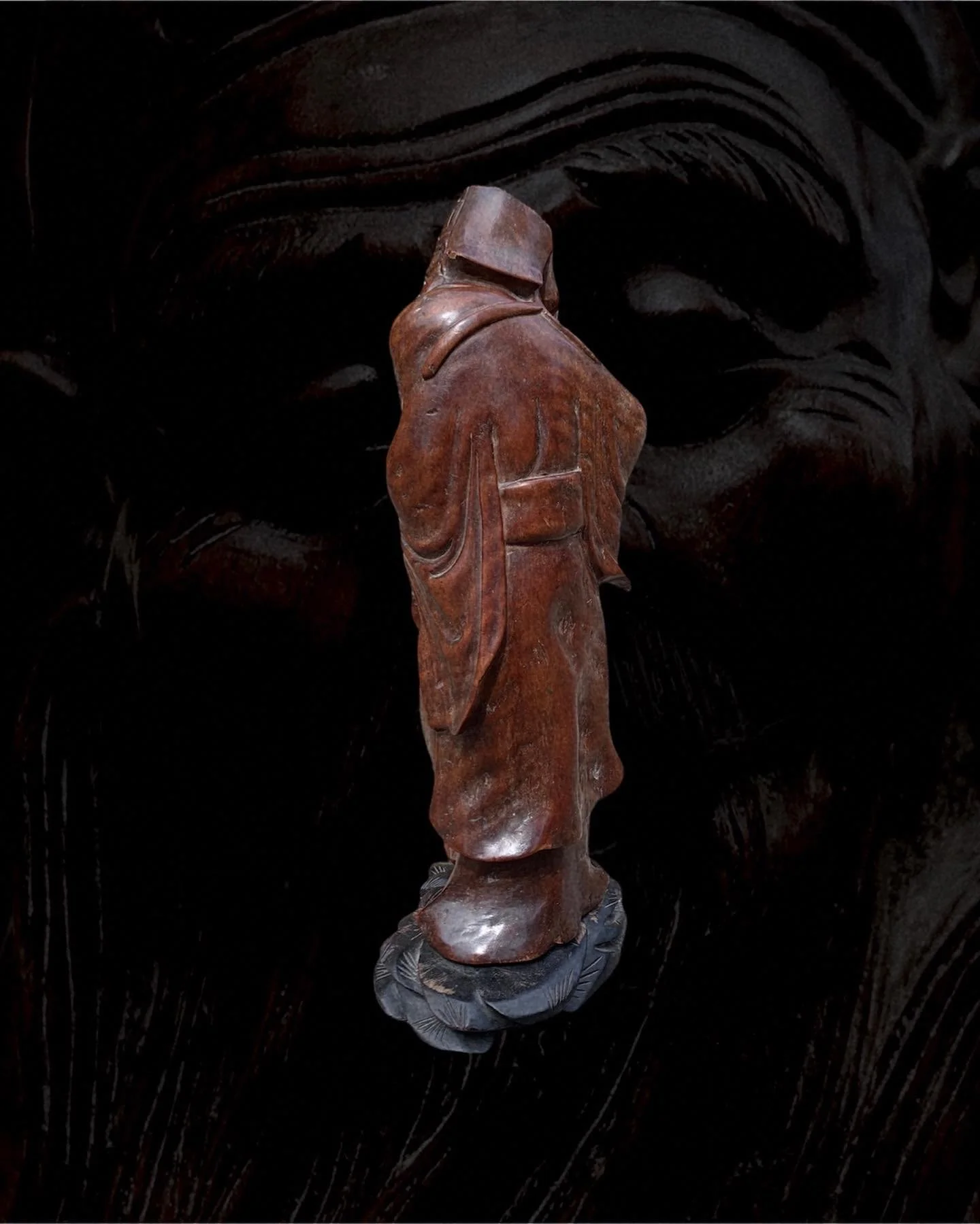A wooden sculpture of a person in traditional attire, without a head visible, standing on a carved wooden platform against a dark background.