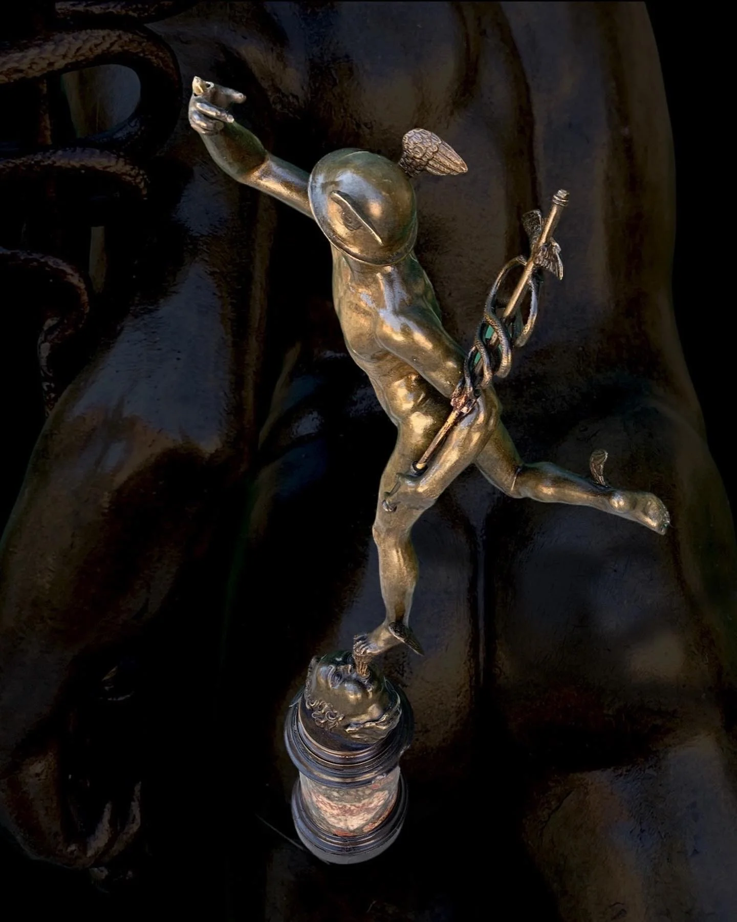 A bronze sculpture of a satyr-like figure with human and goat features, holding a spear and reaching out, mounted on a base shaped like a human face