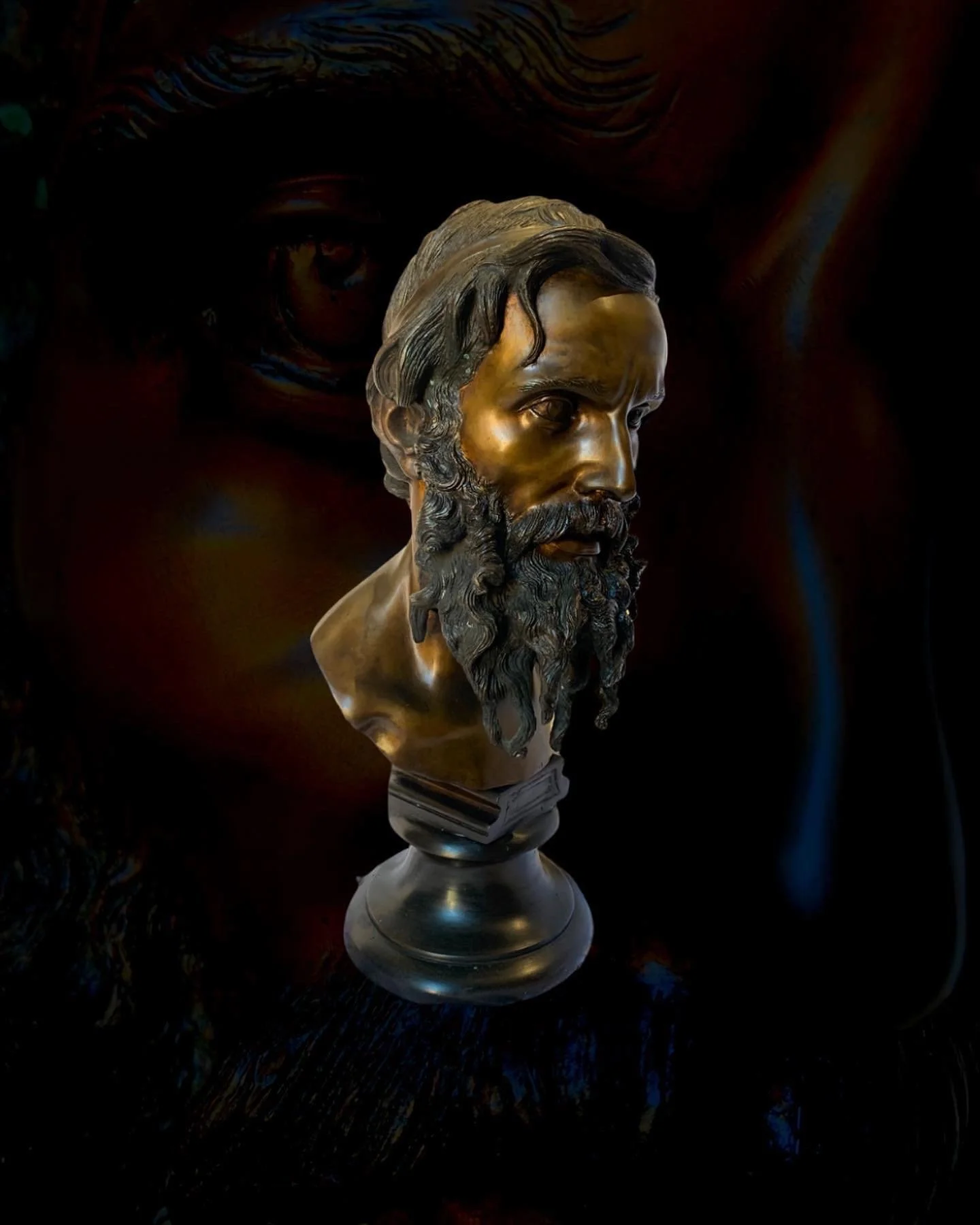 Bronze bust of a bearded man with wavy hair, set against a dark, abstract background.