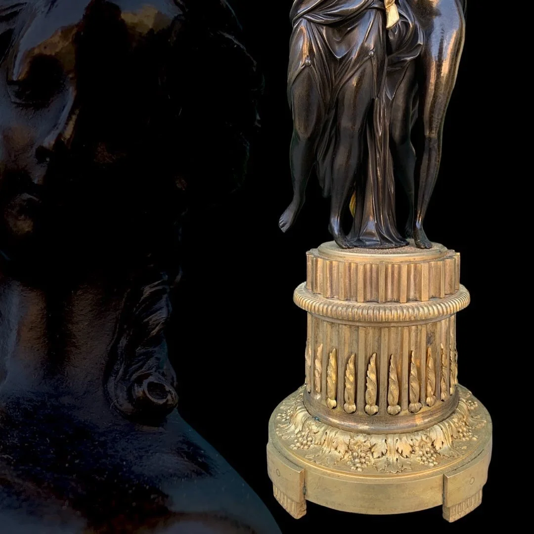 Close-up of an ornate, dark-colored sculpture with a detailed pedestal base, featuring classical design elements, with another similar sculpture partially visible in the background.