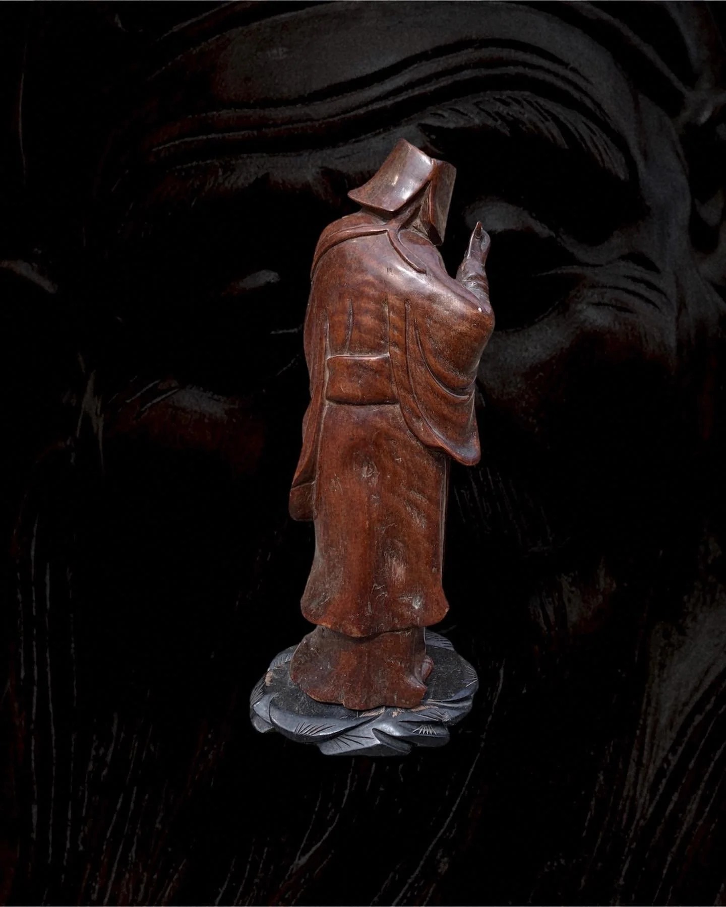 A wooden sculpture of a person standing on a carved black base with a dark, textured background.