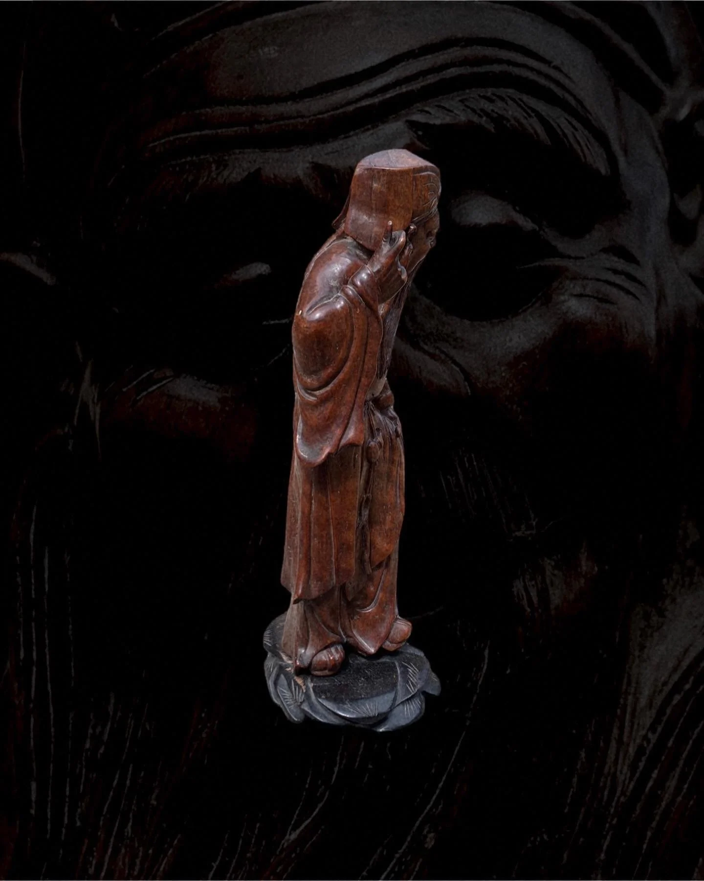 A wooden sculpture of a woman standing on a black base, with her head tilted and hand placed near her ear, set against a dark background with carved designs.