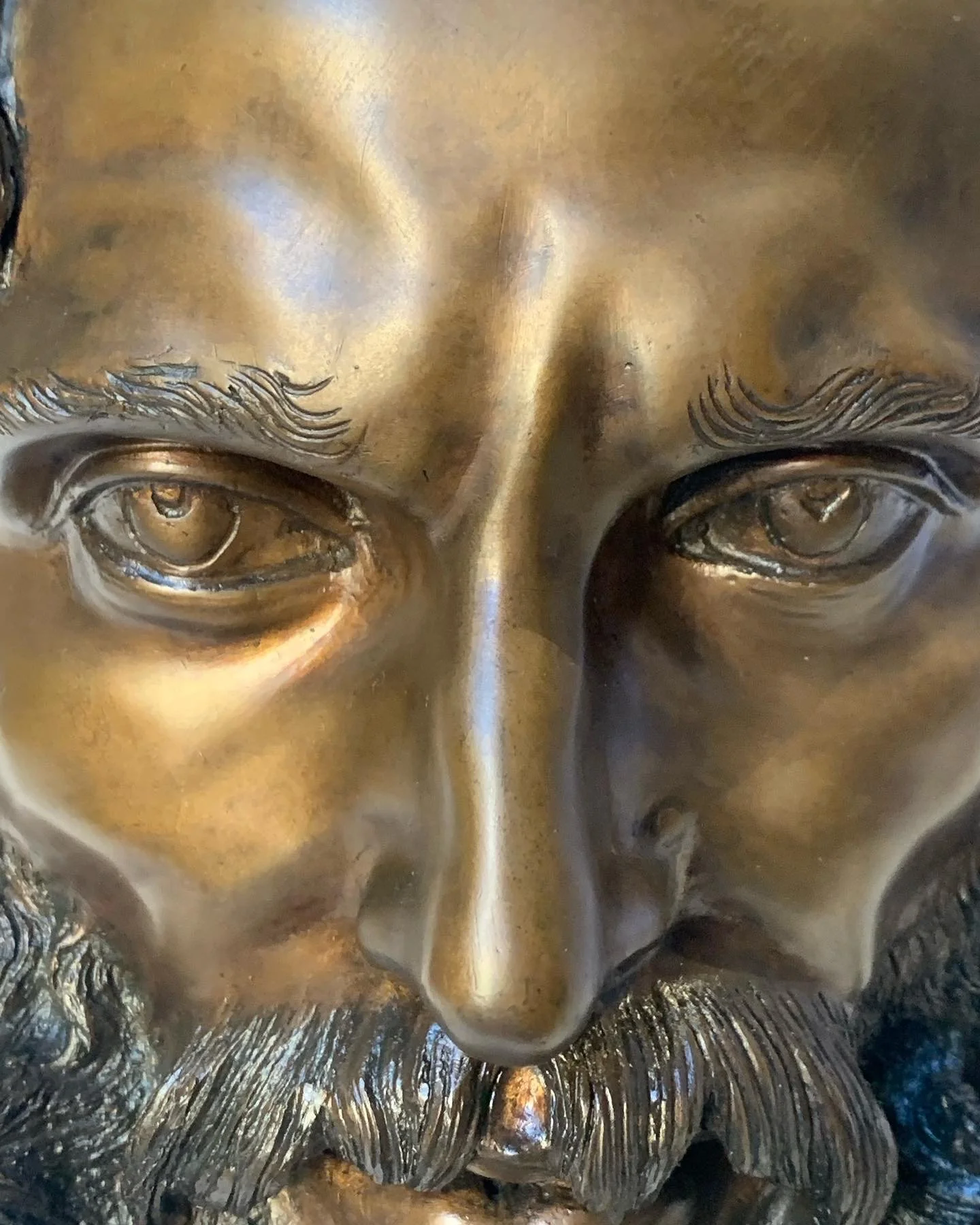 A close-up of a bronze or brass sculpture of a man's face, showing detailed features including eyes, nose, eyebrows, and a full beard.