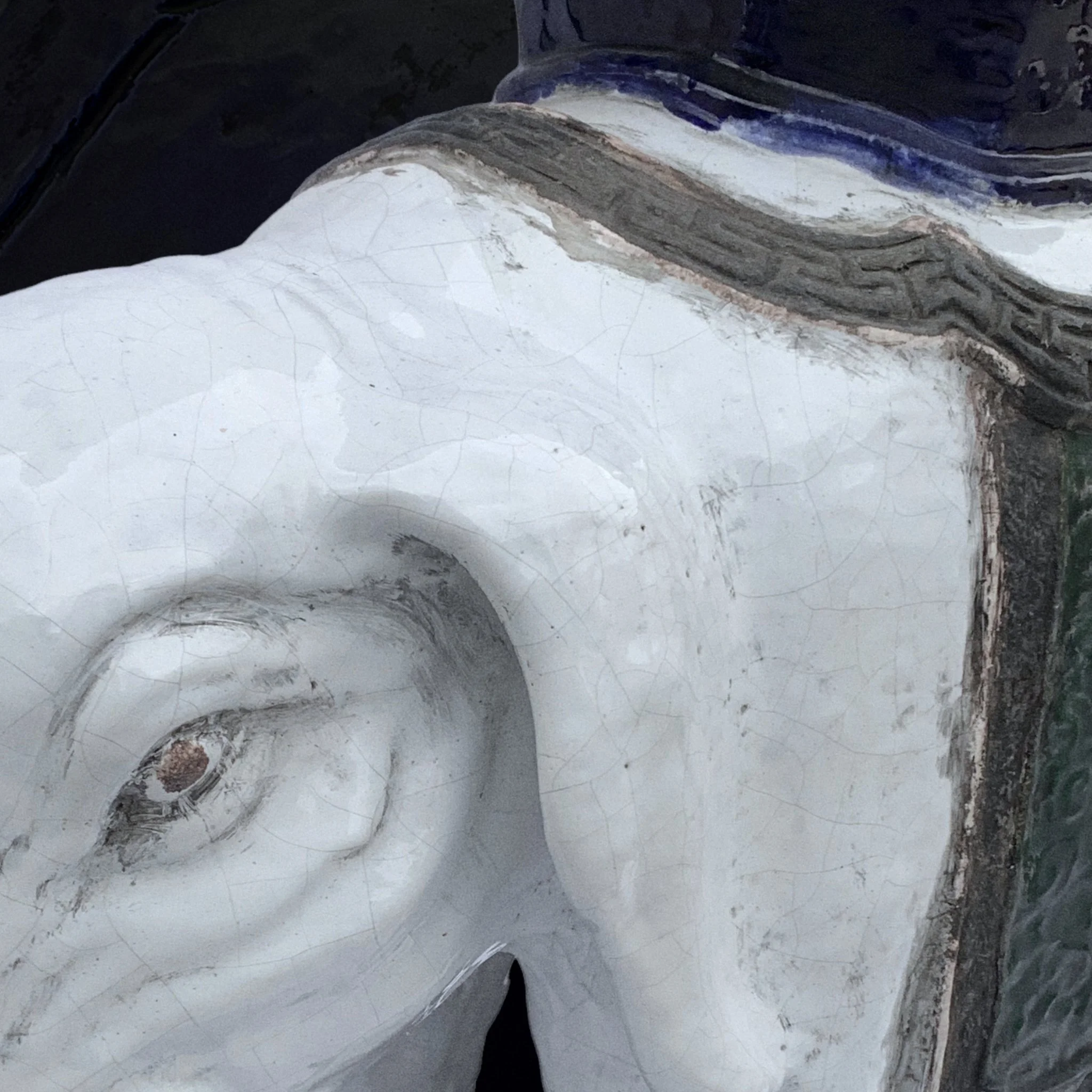 Close-up of a weathered, painted sculpture of a face, showing eye, nose, and part of the head with visible cracks and signs of aging.