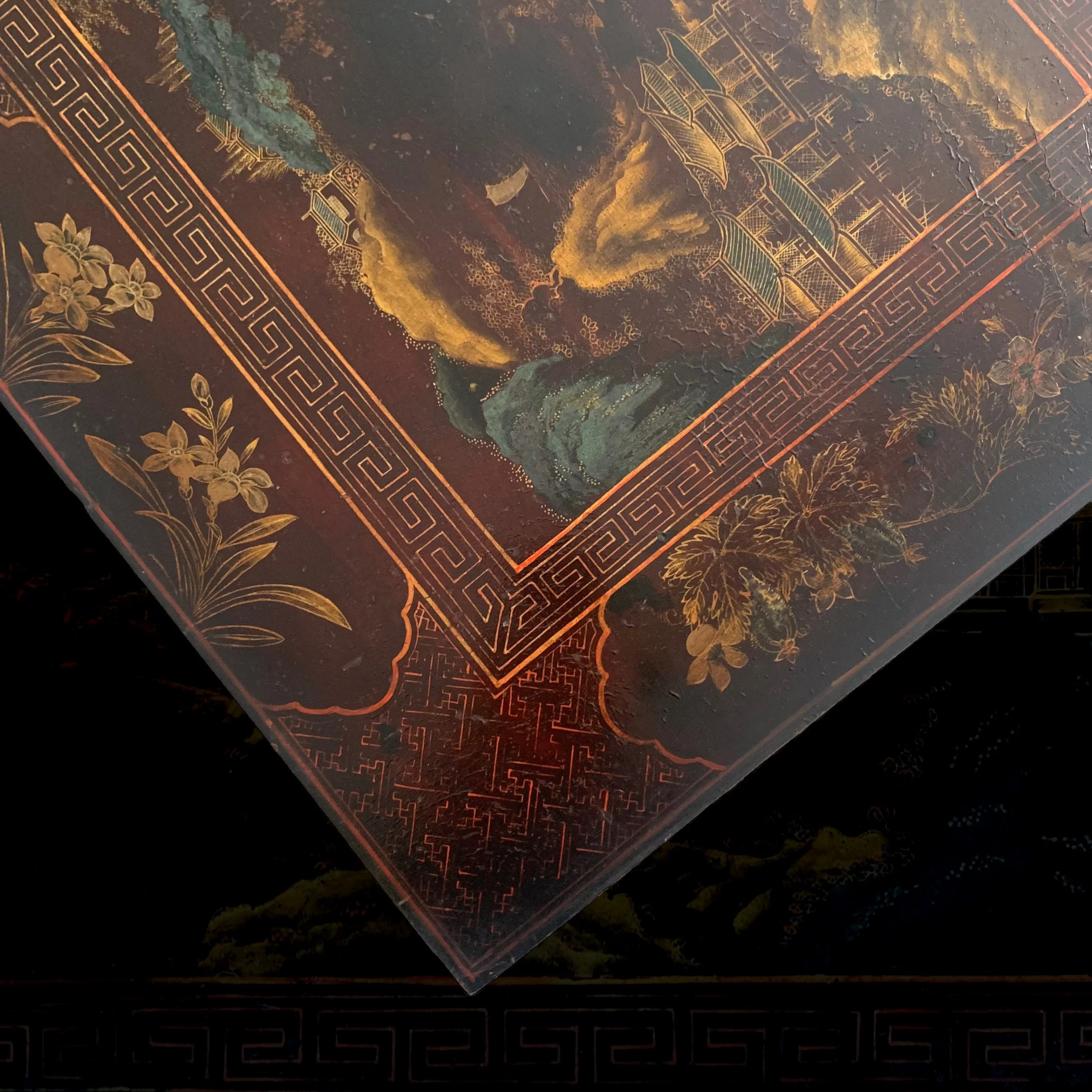 Close-up of an ornate, decorative corner of a traditional Asian-style lacquered box or panel featuring intricate gold and red patterns, floral motifs, and a landscape scene with mountains and a pavilion.