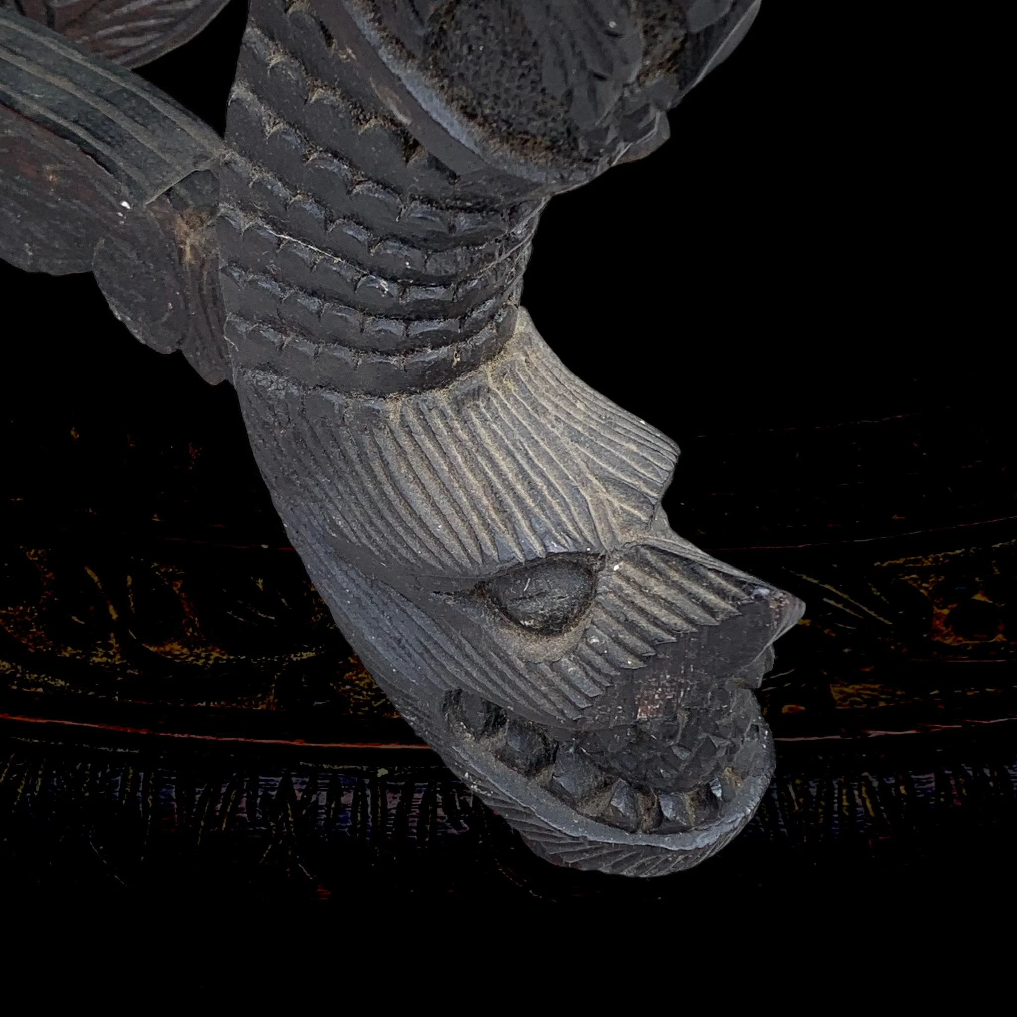 Close-up of a carved wooden figure of a creature with a fierce expression, sharp teeth, and detailed facial features. The figure appears to be part of traditional or indigenous artwork.