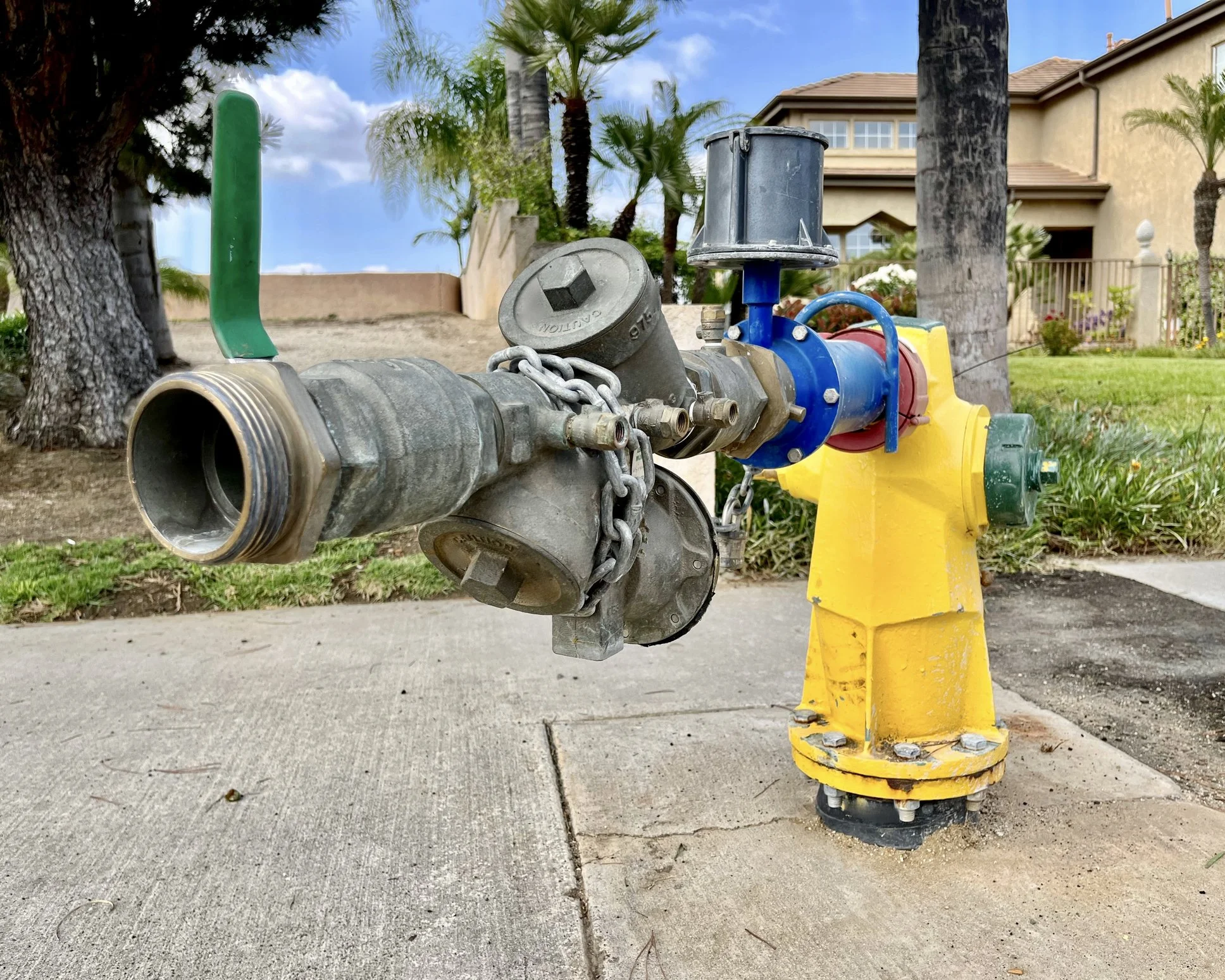 A fire hydrant with a hose and nozzle attached, in a residential neighborhood with trees, grass, and houses in the background.