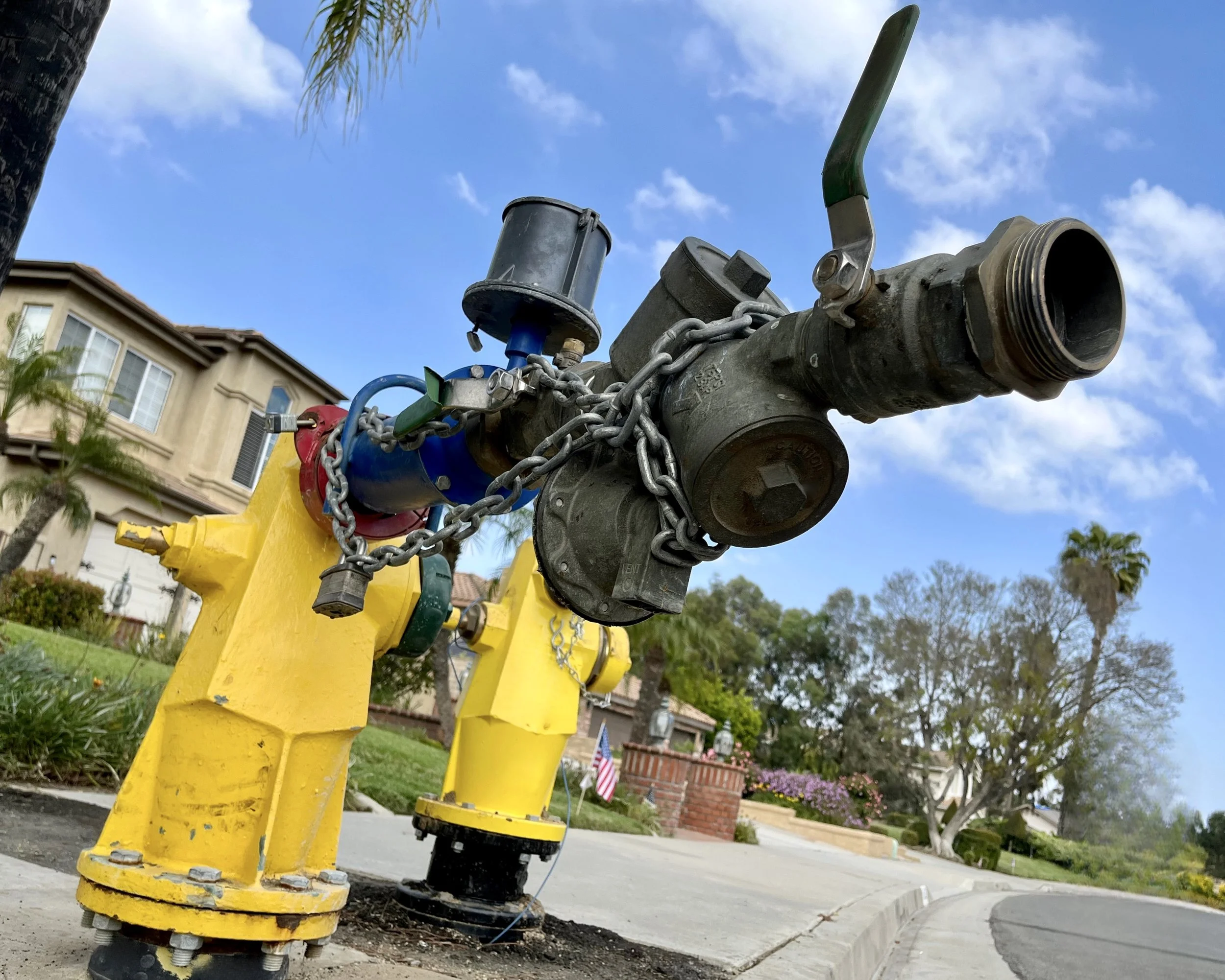 A fire hydrant with a chain attached is installed on a sidewalk in a residential neighborhood, with houses, trees, and a blue sky with some clouds in the background.