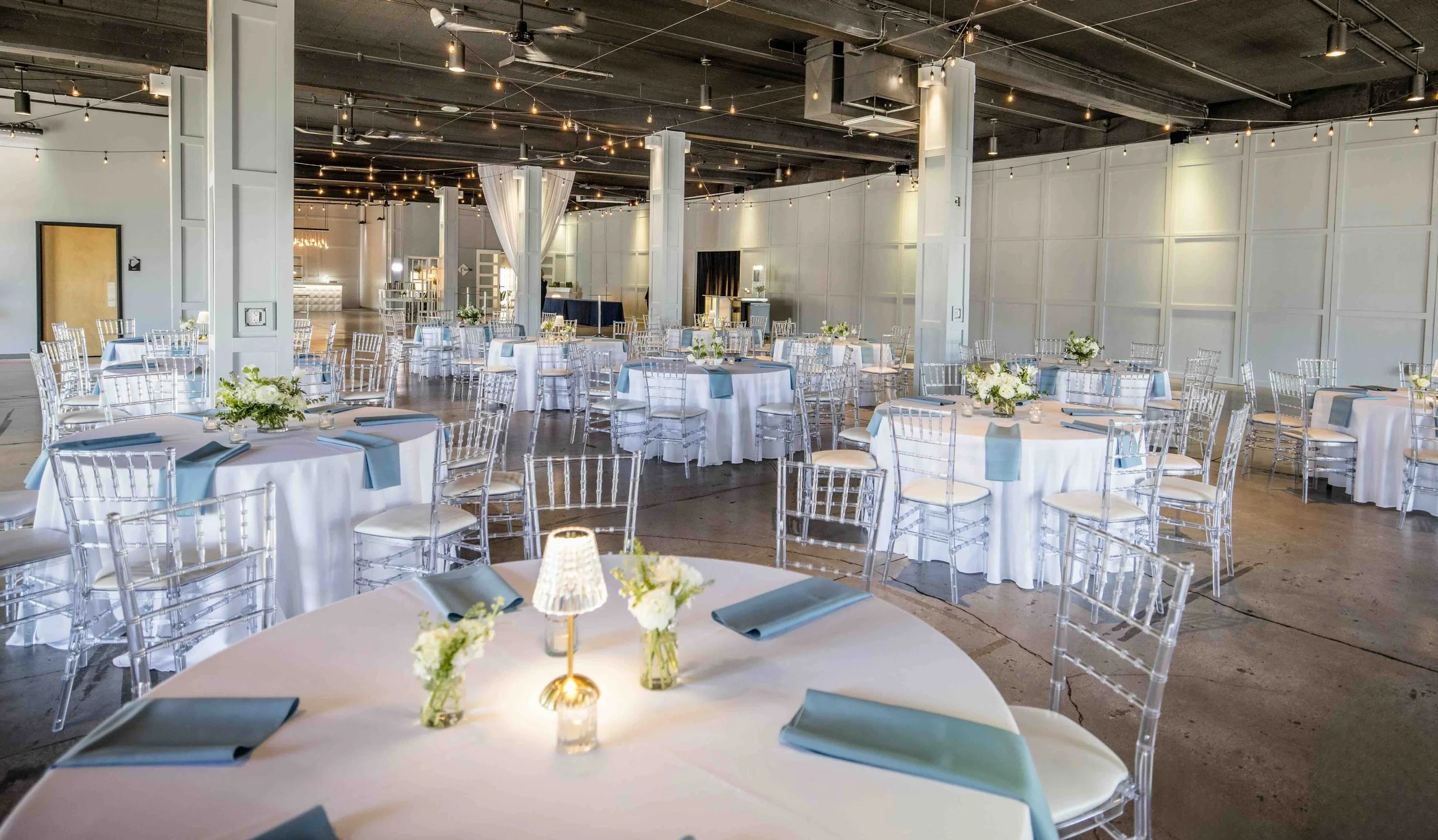 Elegant event space with round tables draped in white tablecloths, decorated with floral centerpieces, blue and white napkins, and small lamps, set up for a formal reception or celebration, featuring clear chairs, string lights overhead, and a spacio