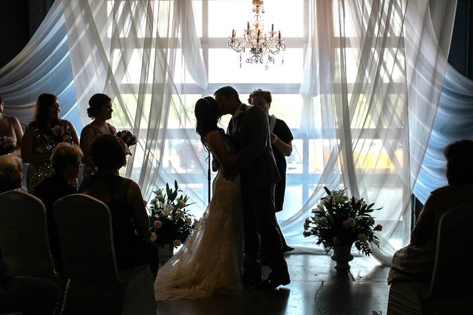 Bride and groom sharing a first kiss during a wedding ceremony, surrounded by seated guests, with large windows and flowing curtains in the background.