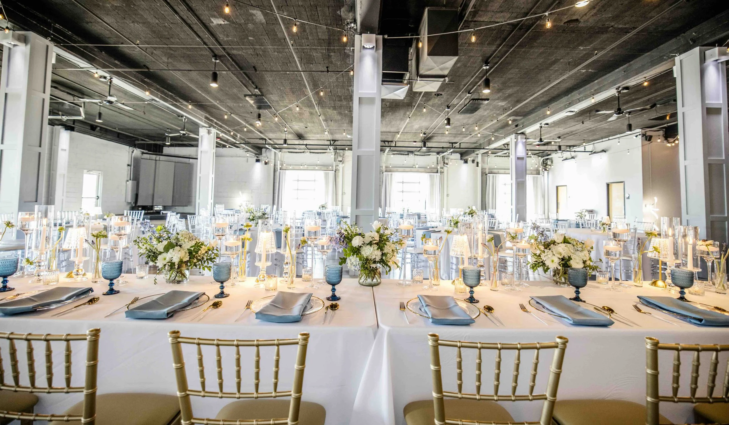 Elegant banquet hall with long tables set with white tablecloths, arranged for a formal event. The tables are decorated with centerpieces of white and green flowers, blue goblets, and gold utensils. Gold Chiavari chairs are positioned around the tabl