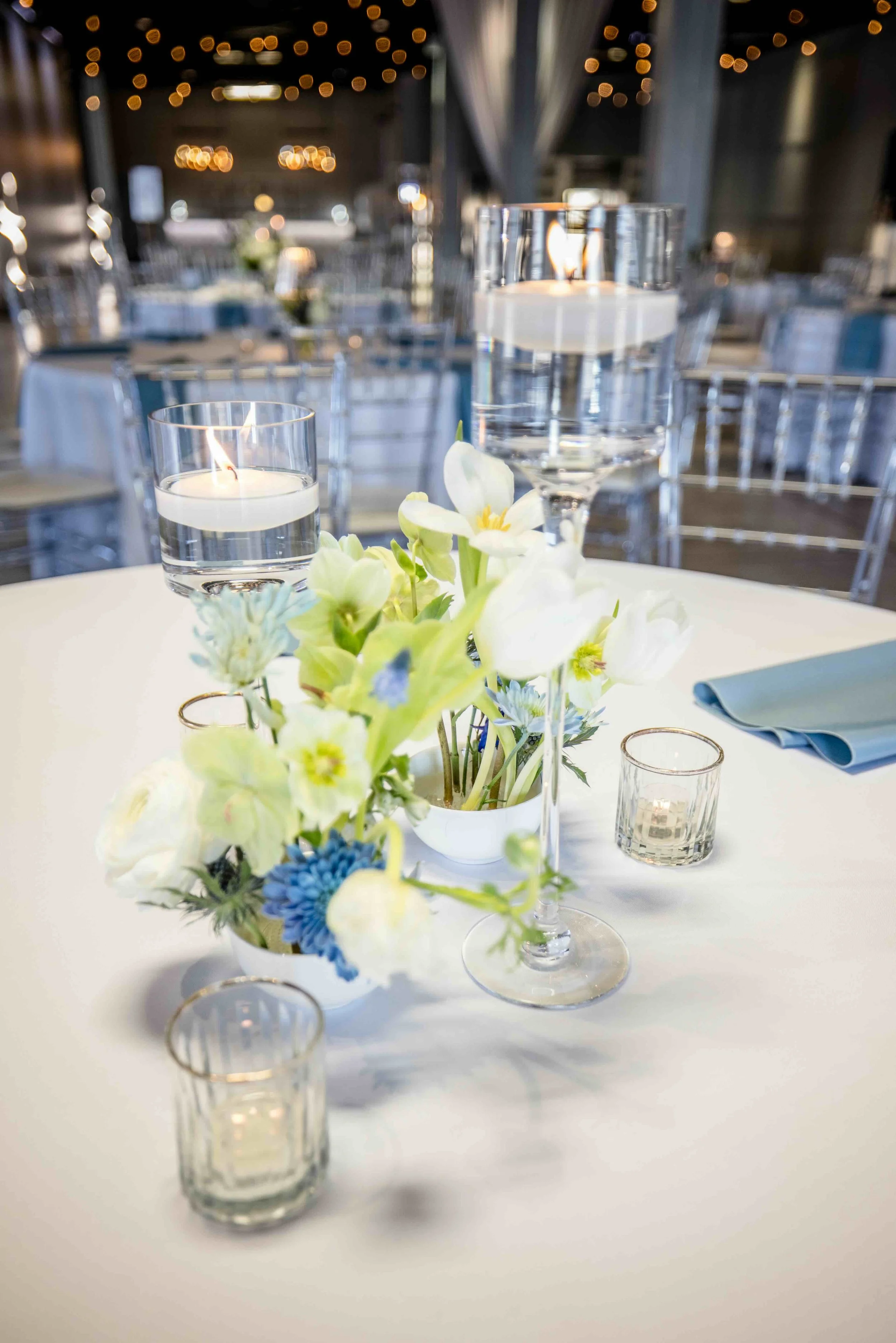 Decorated table at a wedding or event with a floral centerpiece, floating candles, and glassware, with string lights in the background.
