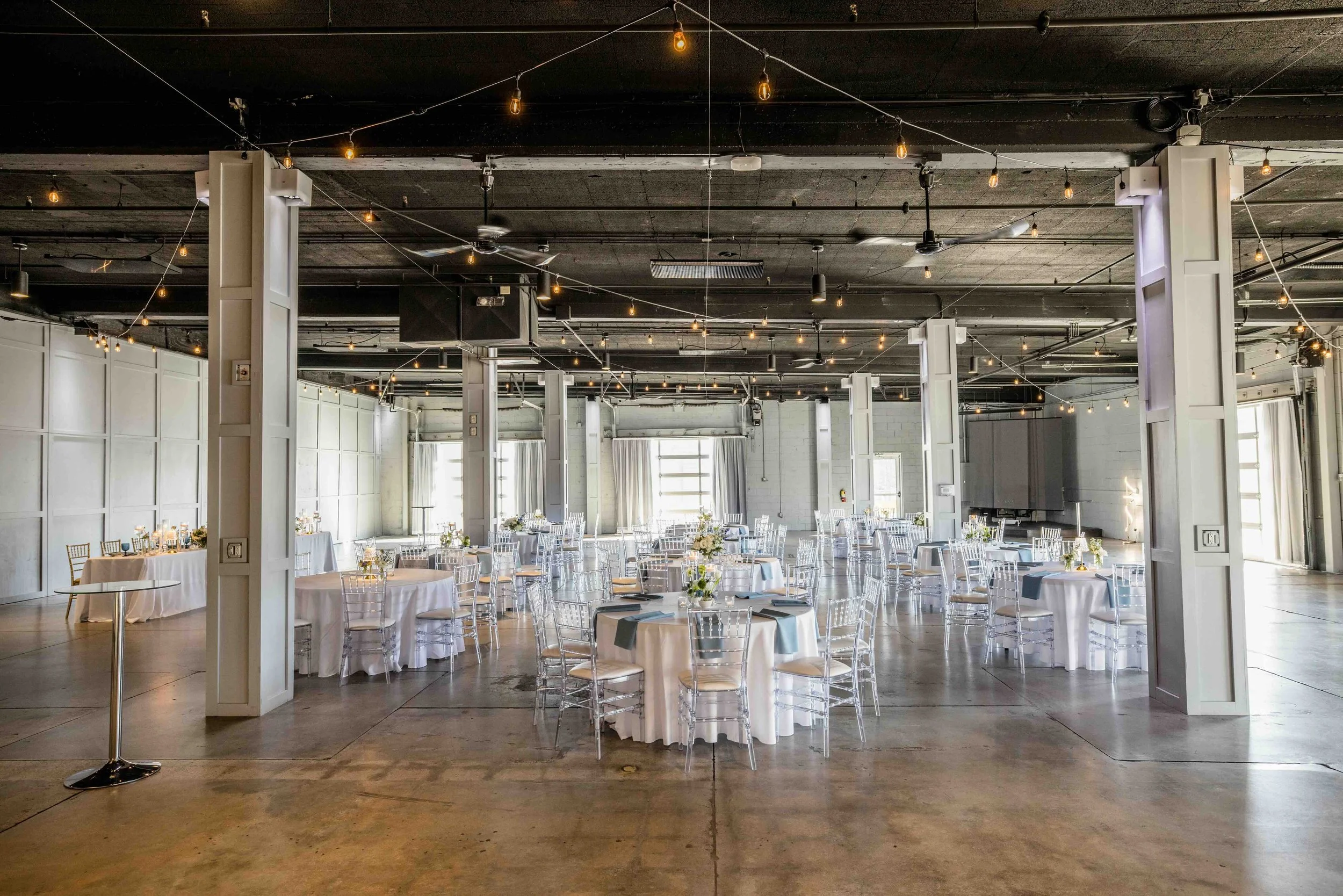 Event space set up with round tables covered in white tablecloths, decorated with floral centerpieces, and surrounded by transparent chairs. String lights decorate the ceiling, and natural light filters through surrounding garage windows.