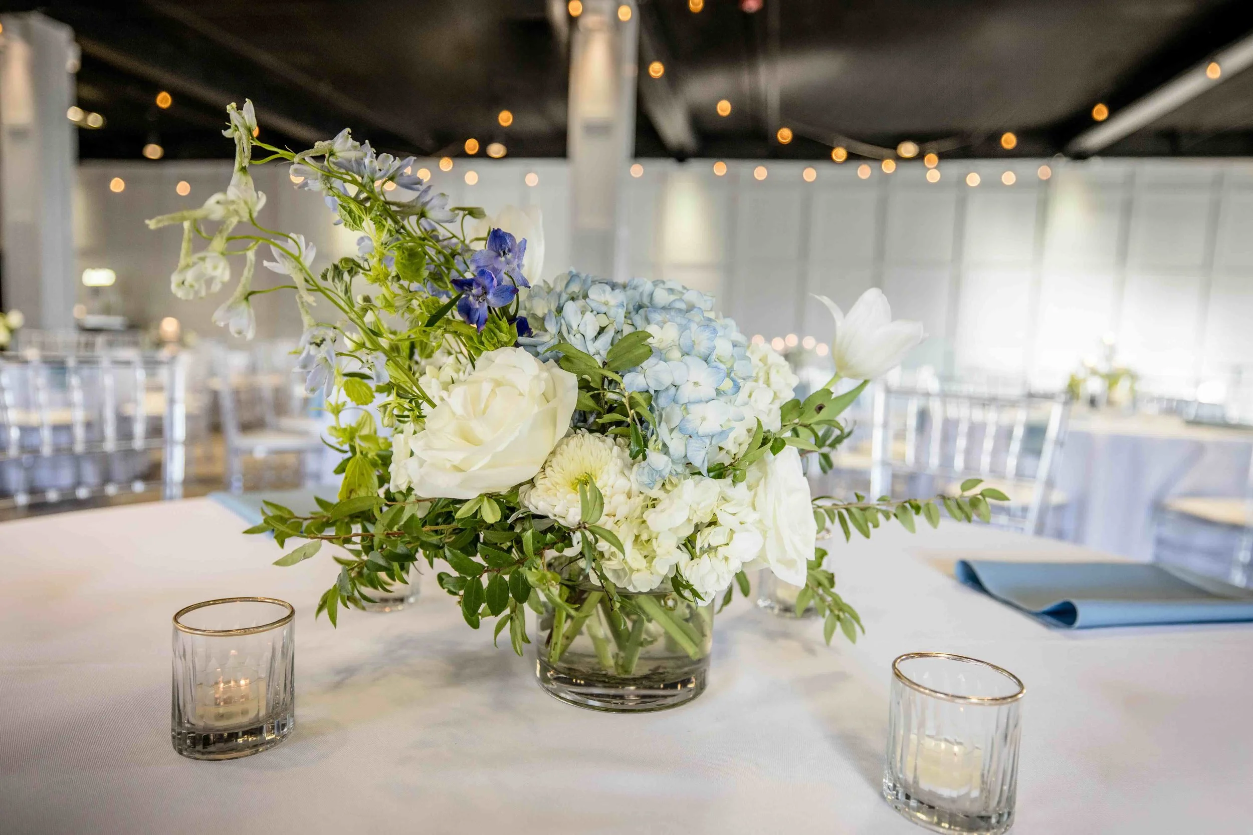Elegant floral centerpiece with white roses, blue hydrangeas, and greenery on a white tablecloth, with candle holders and blue napkin, in a decorated event hall with string lights.