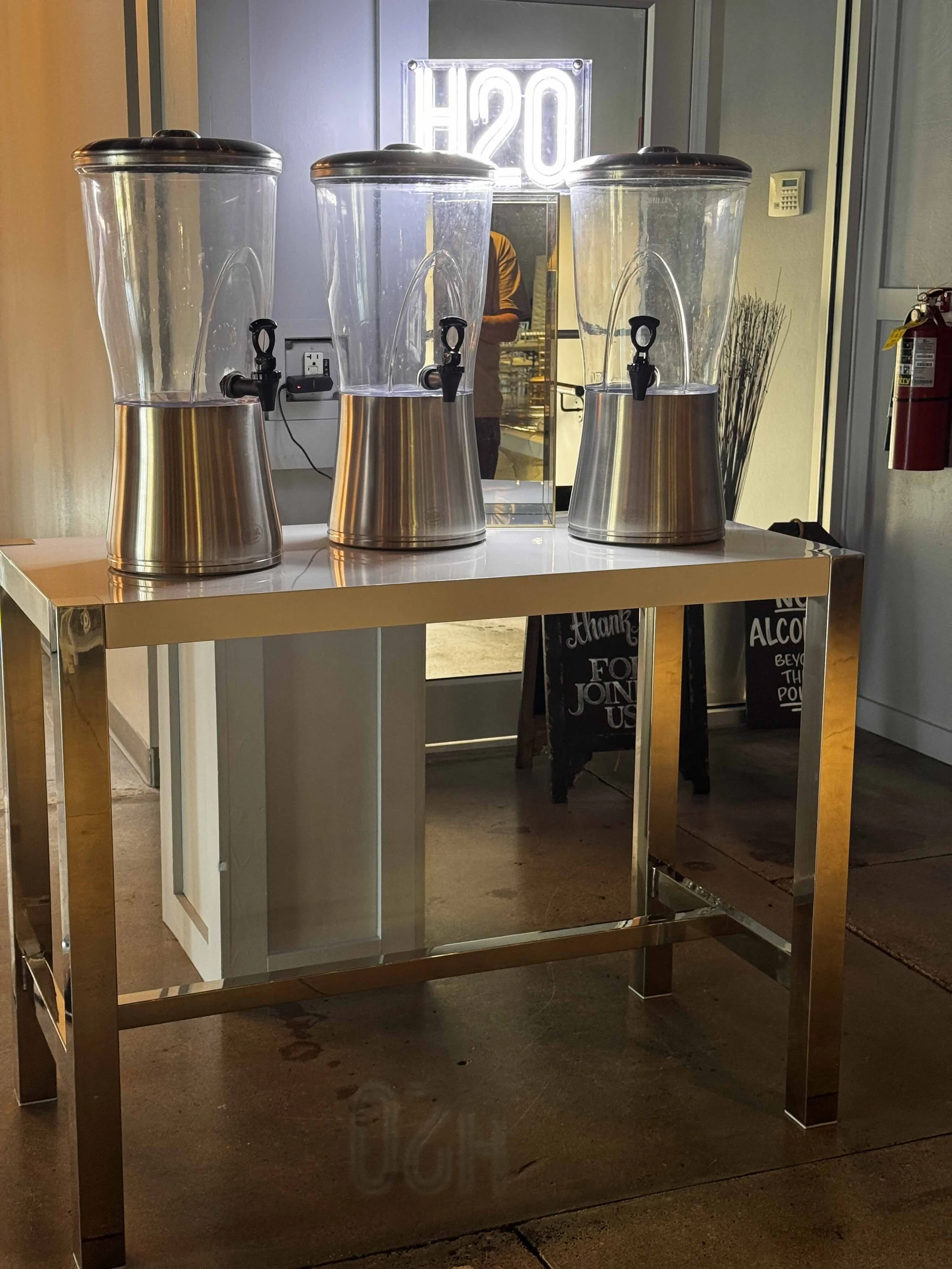 Three large transparent dispensers with spigots on a white metal table, with a neon 'H2O' sign representing a premium water service.