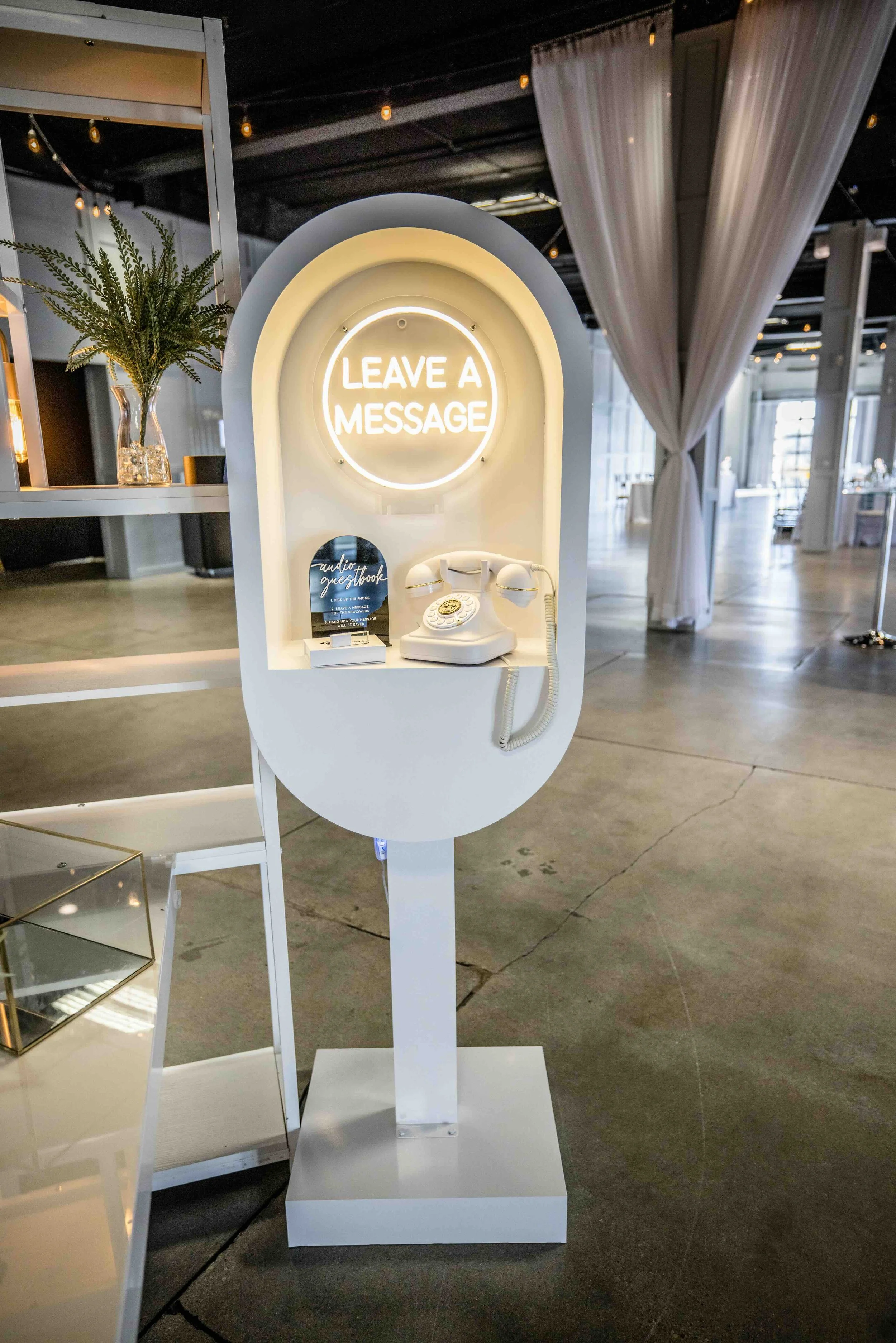 A white photo booth with a neon sign that says "Leave a Message," a vintage rotary phone, and a guestbook. The booth is in a spacious room with string lights, white curtains, and decorative plants.