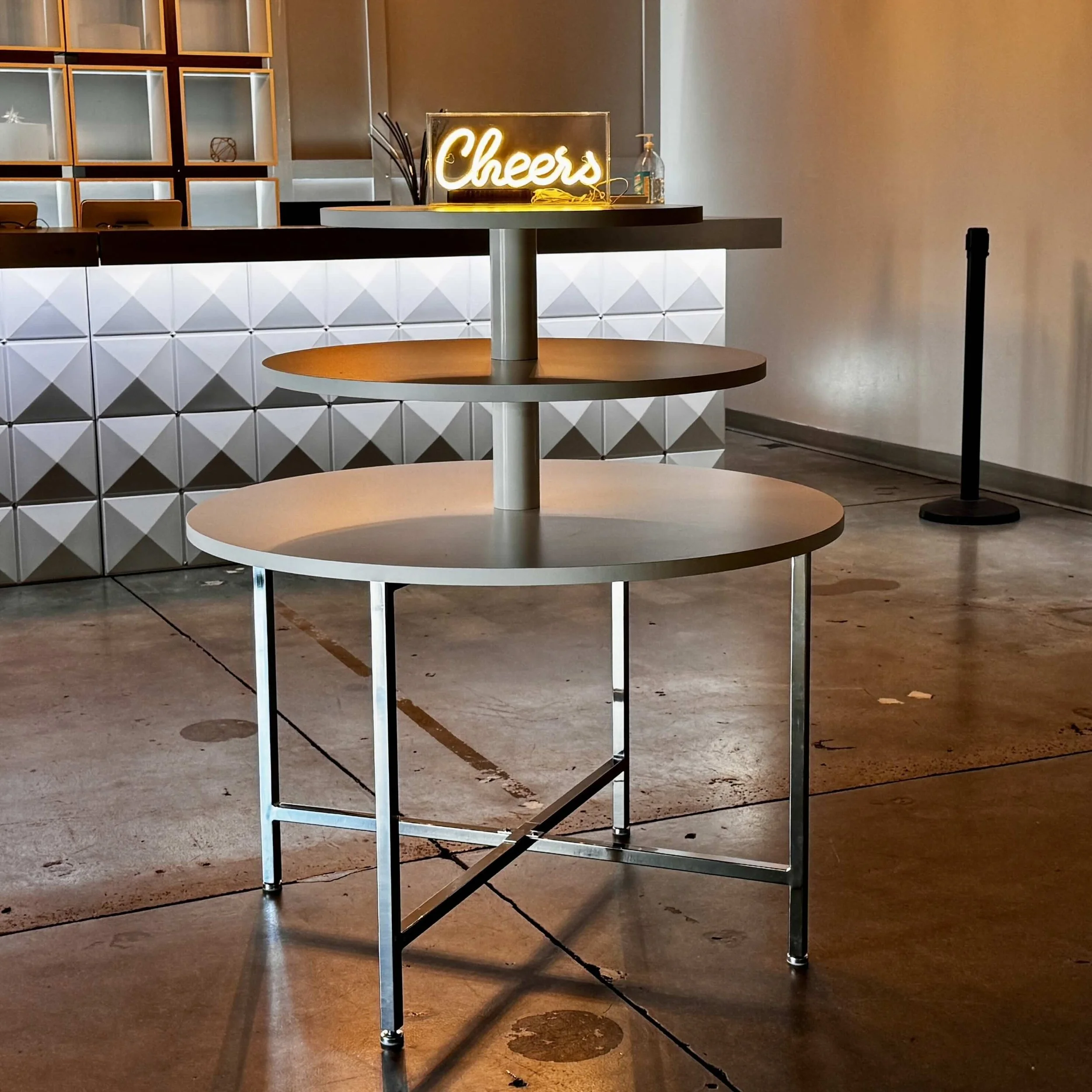 A round, tiered table standing on a polished concrete floor in a modern, brightly lit indoor setting with a neon 'Cheers' sign on the counter behind it, used for serving welcome drinks to guests.