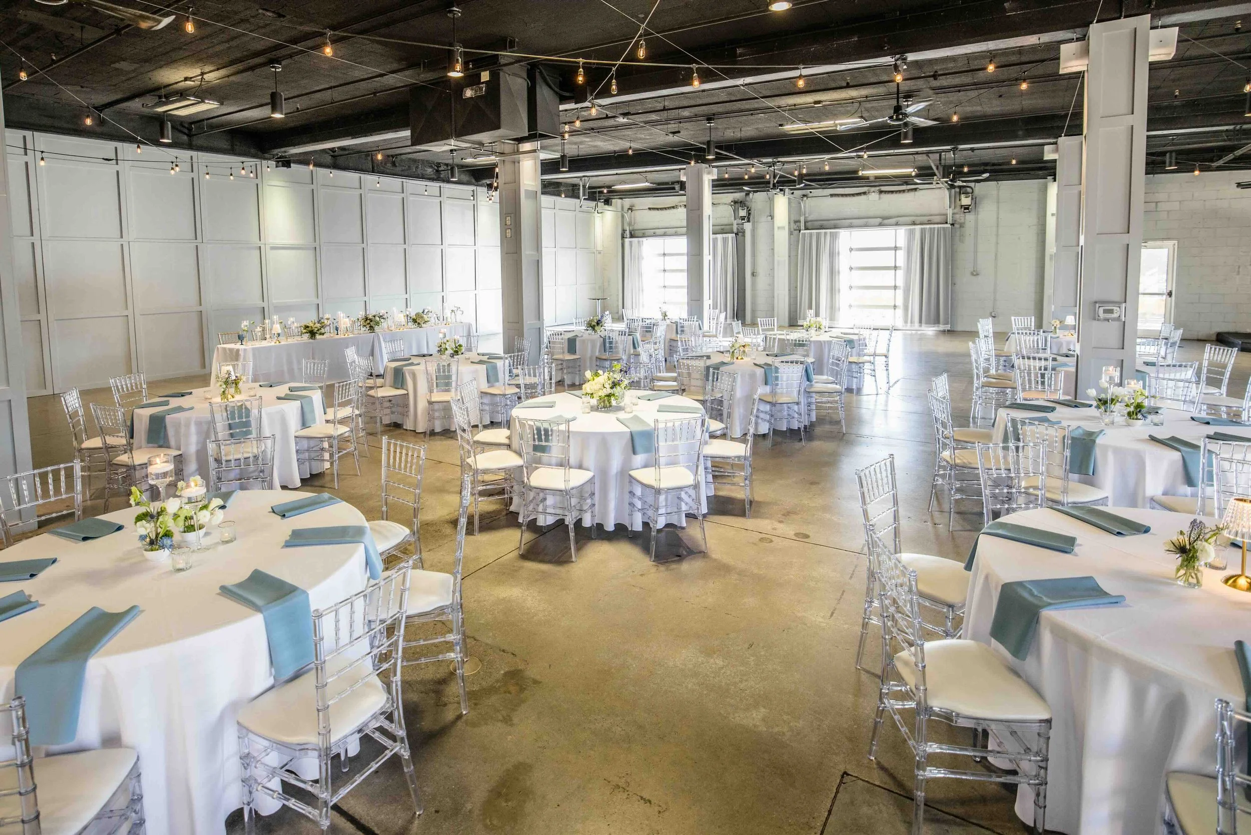 Elegant event space with round tables covered in white tablecloths, adorned with small floral centerpieces and teal napkins. Clear chairs surround the tables, and a long table with floral arrangements runs along the back wall. The room has string lig