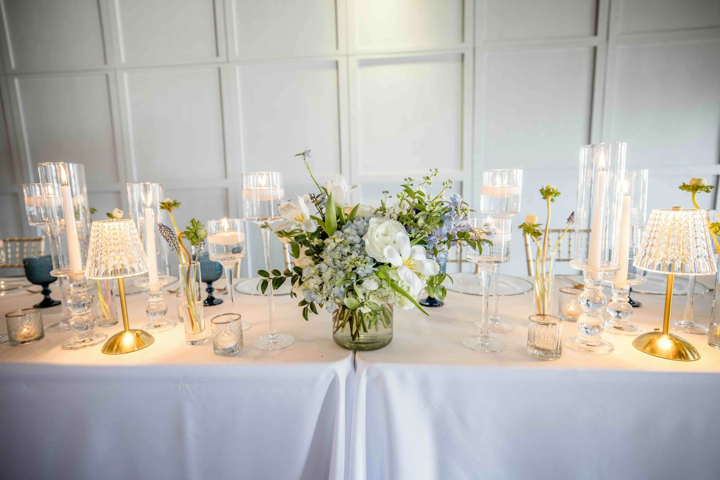 Elegant wedding reception table decorated with white and blue floral centerpiece, glass candle holders, small table lamps with gold bases, and floating candles in tall glass vases.