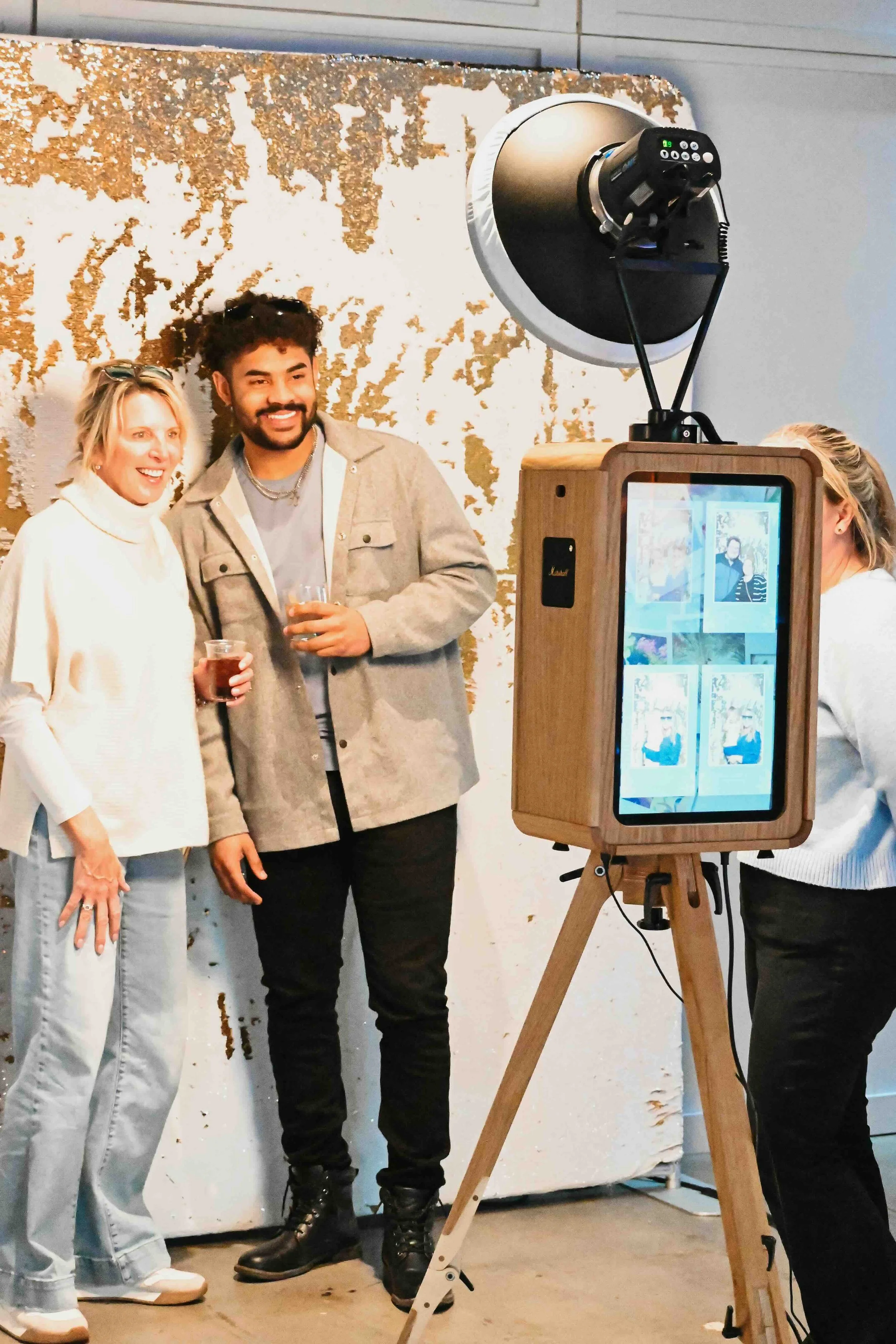Two women and one man smiling and posing for photos with drinks in their hands, being photographed in front of a textured gold and white wall, with a photographer capturing the moment using a vintage-style photo booth.