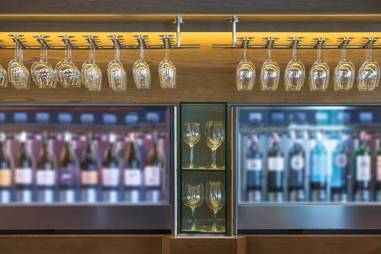 Glassware including upside-down wine glasses hanging upside down from a rack above and empty wine glasses below, with wine bottles behind glass in a bar setting.