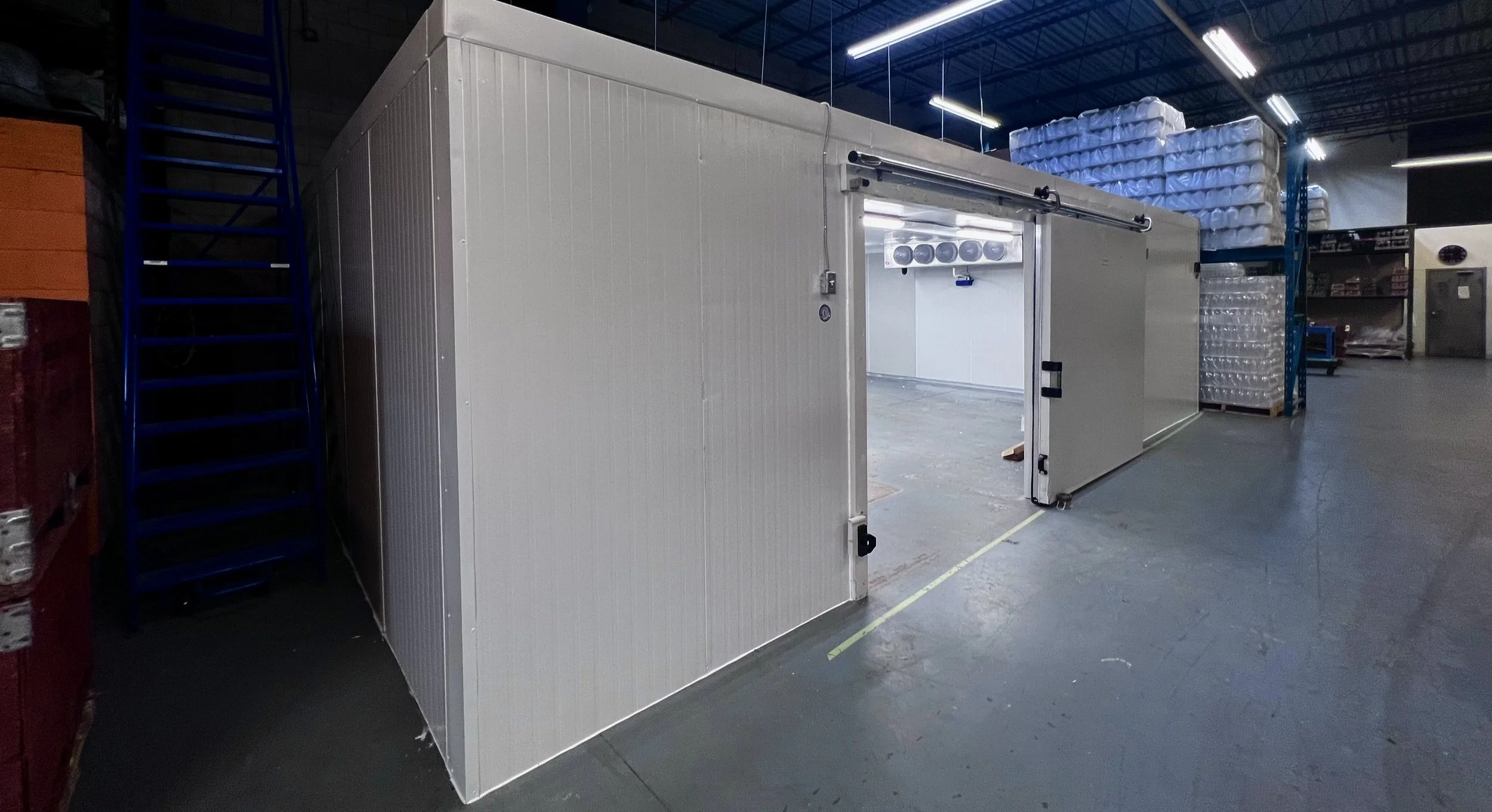 An industrial cold storage room or walk-in freezer inside a warehouse, with metal walls, a sliding door, and overhead lighting.