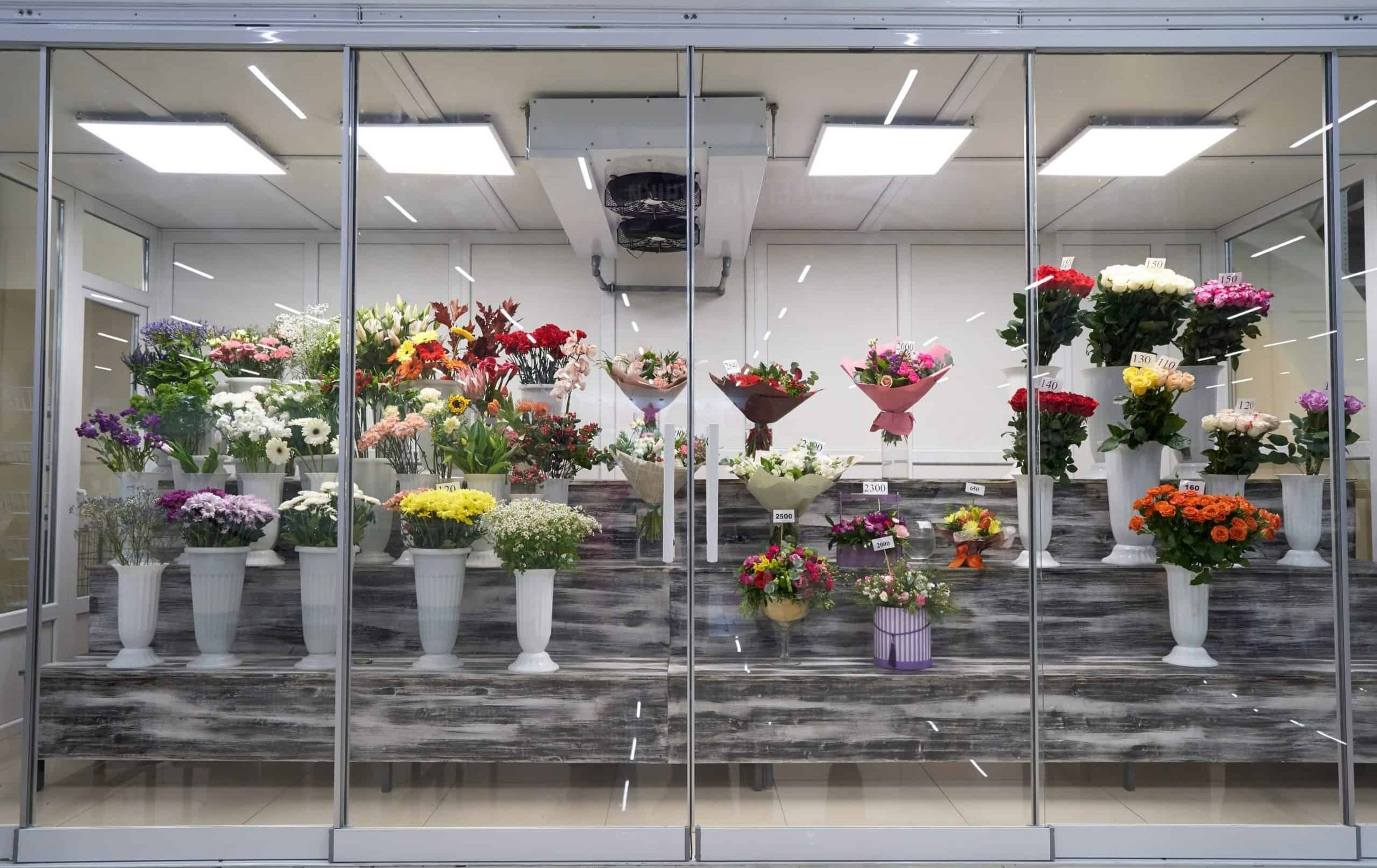 A display of various colorful flowers in vases inside a flower shop. The flowers include lilies, roses, daisies, and others, arranged on a tiered wooden stand behind glass doors.