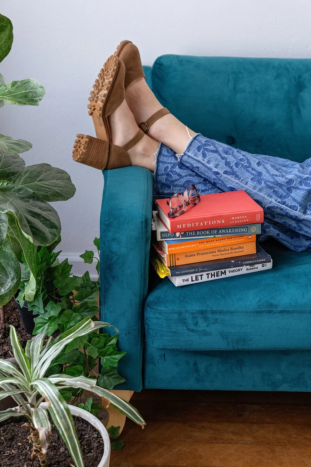 Person with feet up on a teal sofa, wearing brown sandals and blue embroidered jeans, beside a stack of books and potted plants.