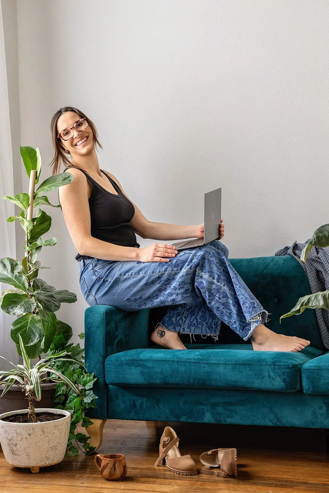 A woman sitting on the back of a teal sofa, holding a silver laptop, smiling at the camera, wearing glasses, a black tank top, and blue patterned jeans. There are potted plants, shoes, and a cup on the wooden floor nearby.