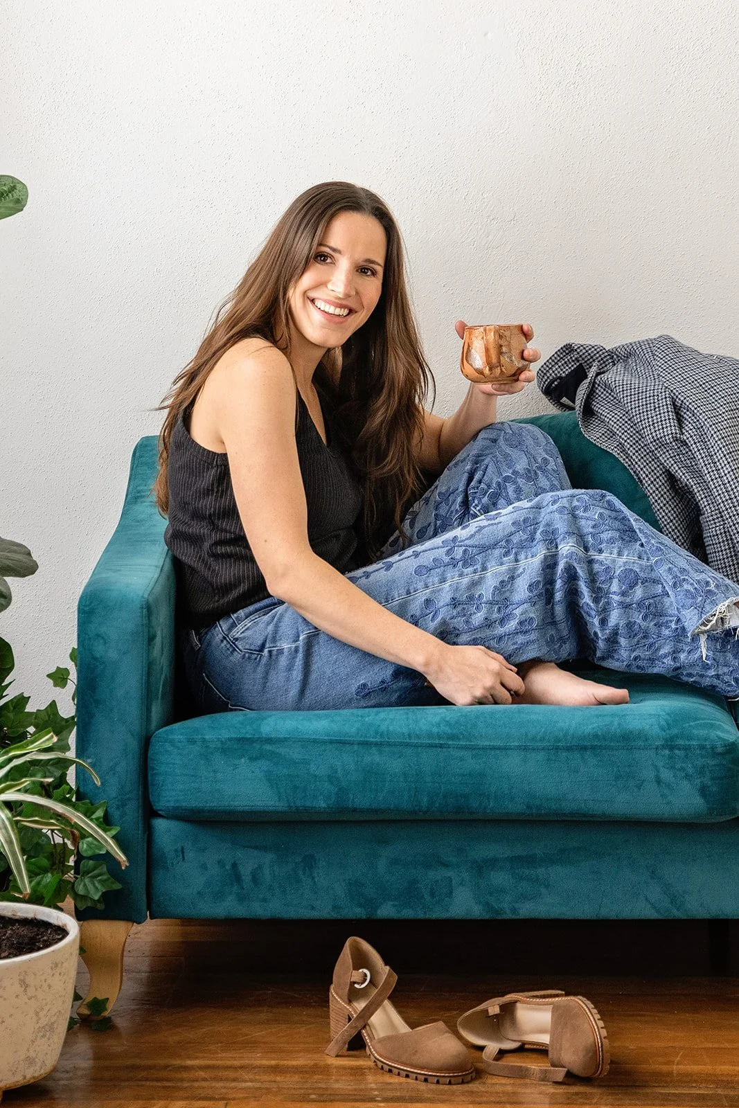 A young woman with long brown hair smiling, sitting on a teal sofa, wearing a black sleeveless top and blue embroidered jeans, holding a ceramic mug, with shoes on the wooden floor in front of her, and a plant nearby.