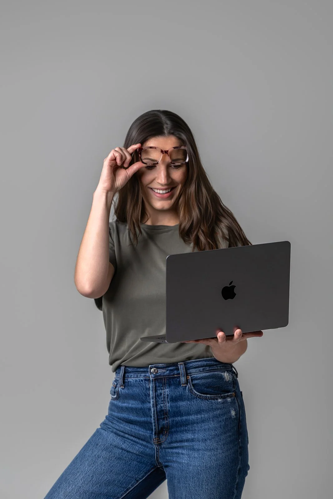 Young woman with long brown hair adjusting her glasses while looking at a silver Apple MacBook against a plain gray background.
