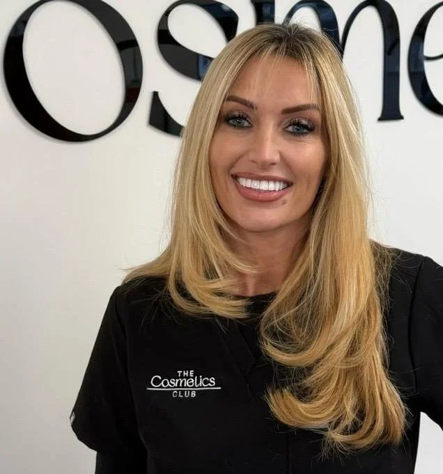 A smiling woman with long blonde hair wearing a black T-shirt that reads 'The Cosmetics Club' standing in front of a white wall with part of a large black logo.