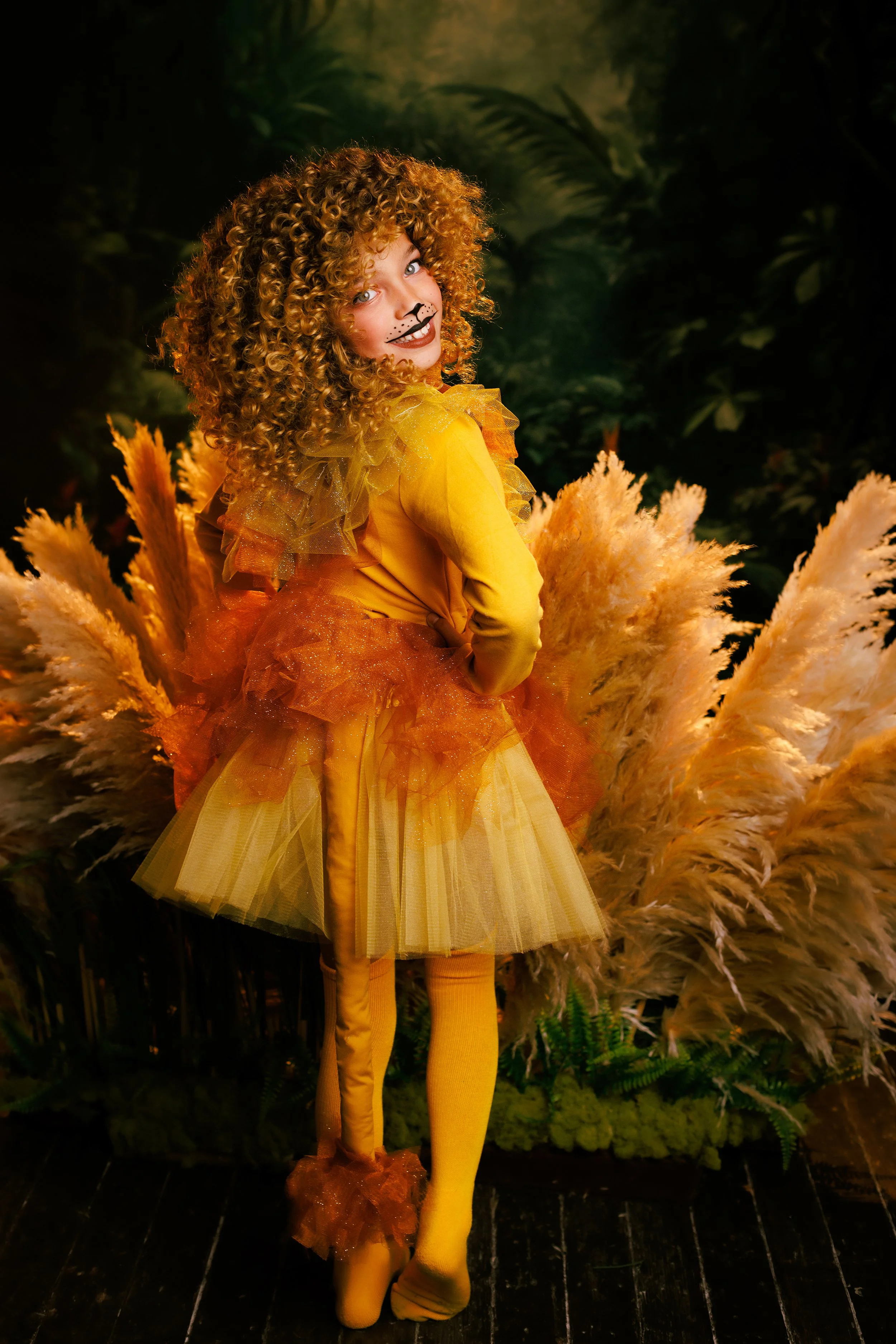 Child dressed as a lion with a yellow and orange costume, curly hair, and face paint, standing in front of pampas grass and green foliage.