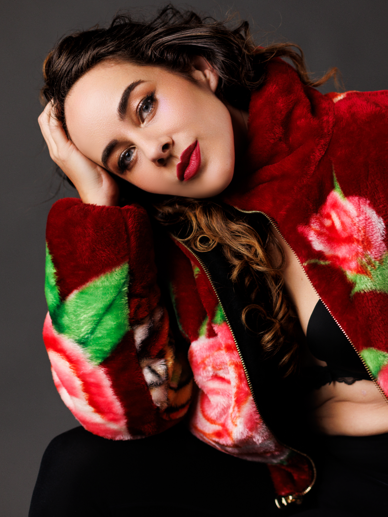 A woman with dark wavy hair and red lipstick wearing a floral patterned red and pink fuzzy jacket, resting her head on her hand against a dark background.